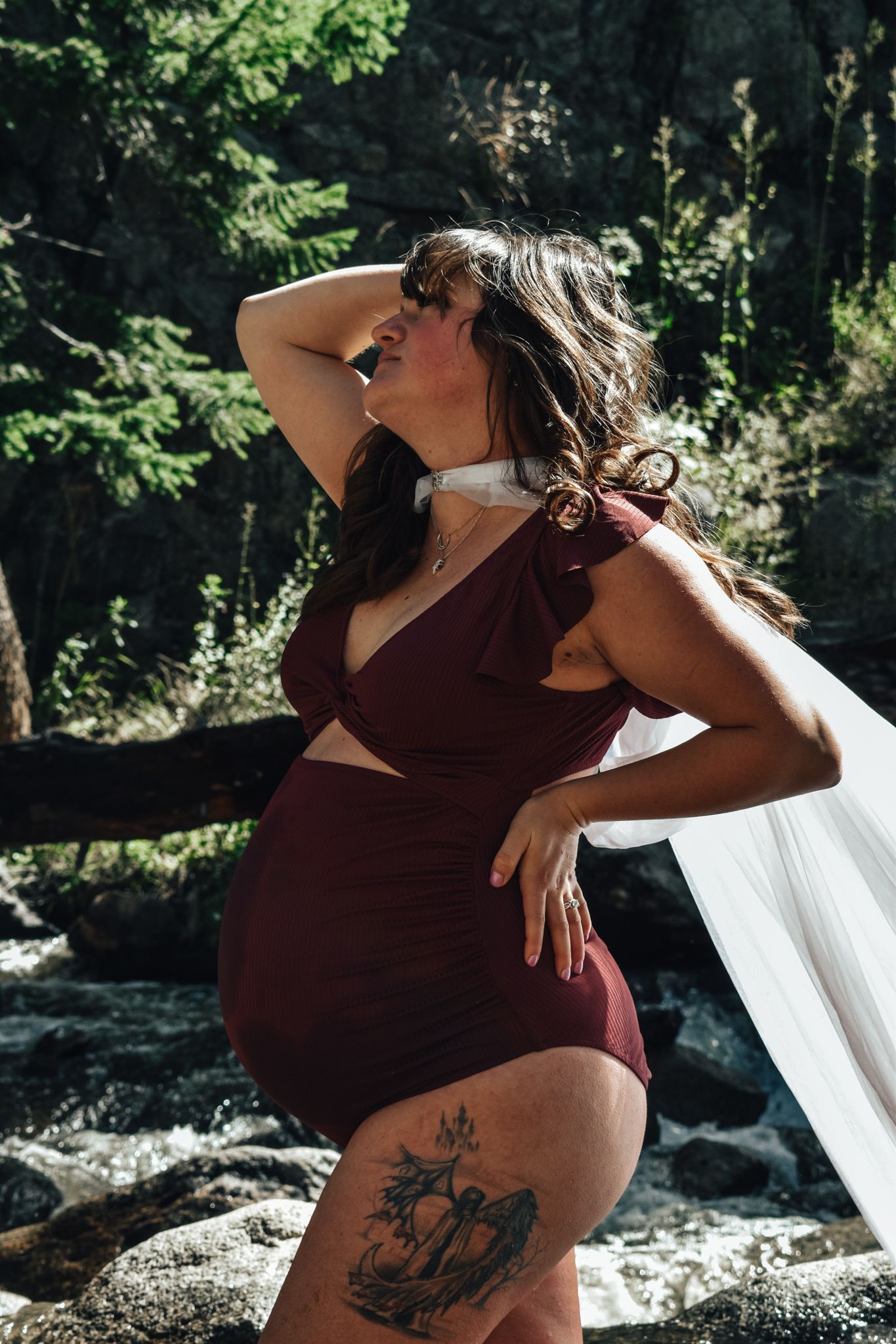 Pregnant woman in burgundy maternity outfit with thigh tattoo posing by a forest waterfall. Maternity Photographer in Pflugerville, Tx