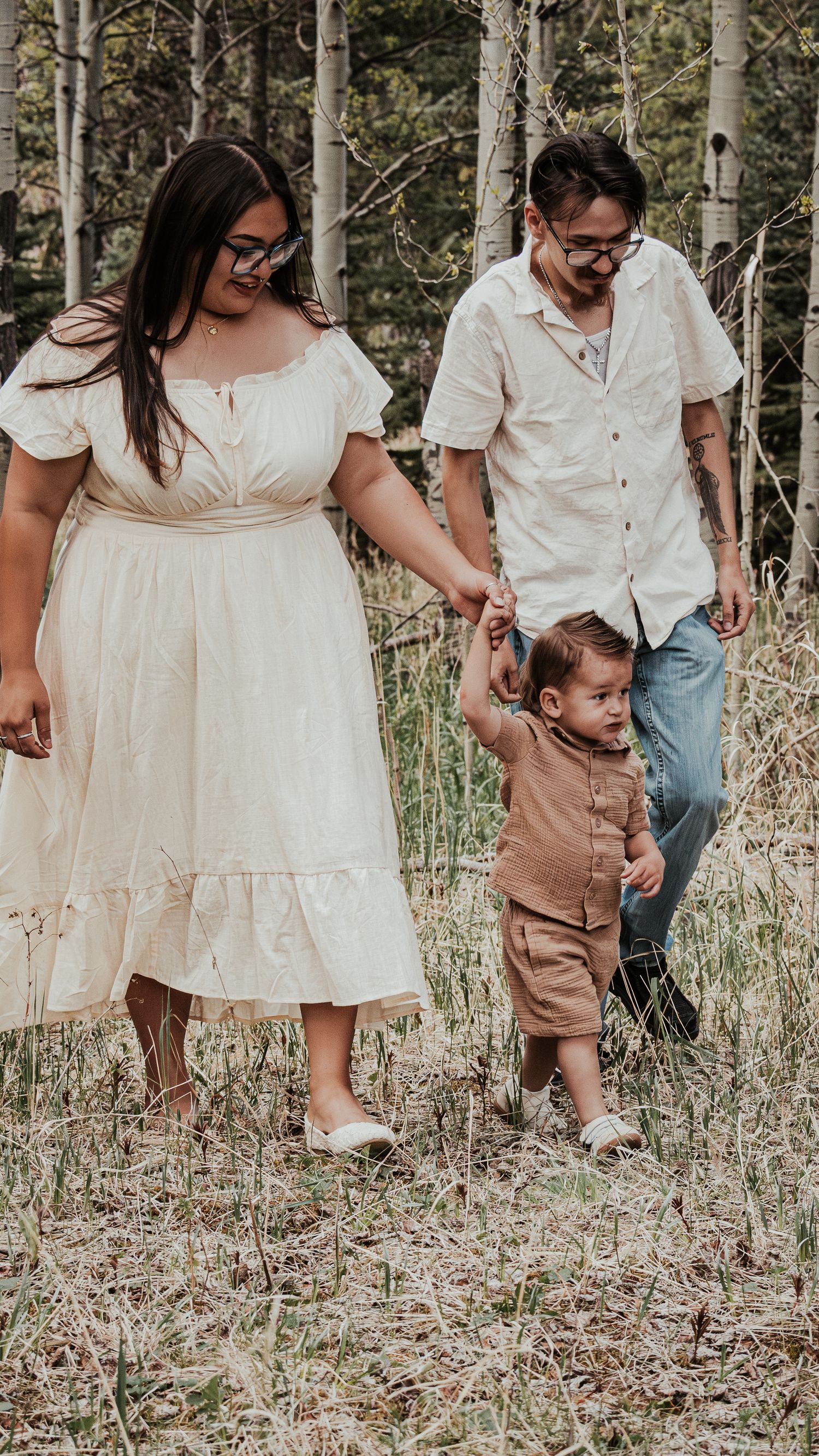 Young family walking together in nature, parents holding toddler's hands among tall grass and trees.