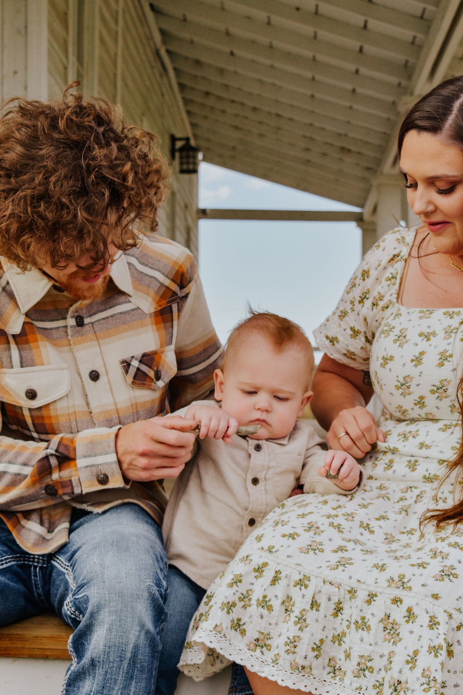 Young parents in plaid and floral outfits sitting with baby on wooden porch steps.