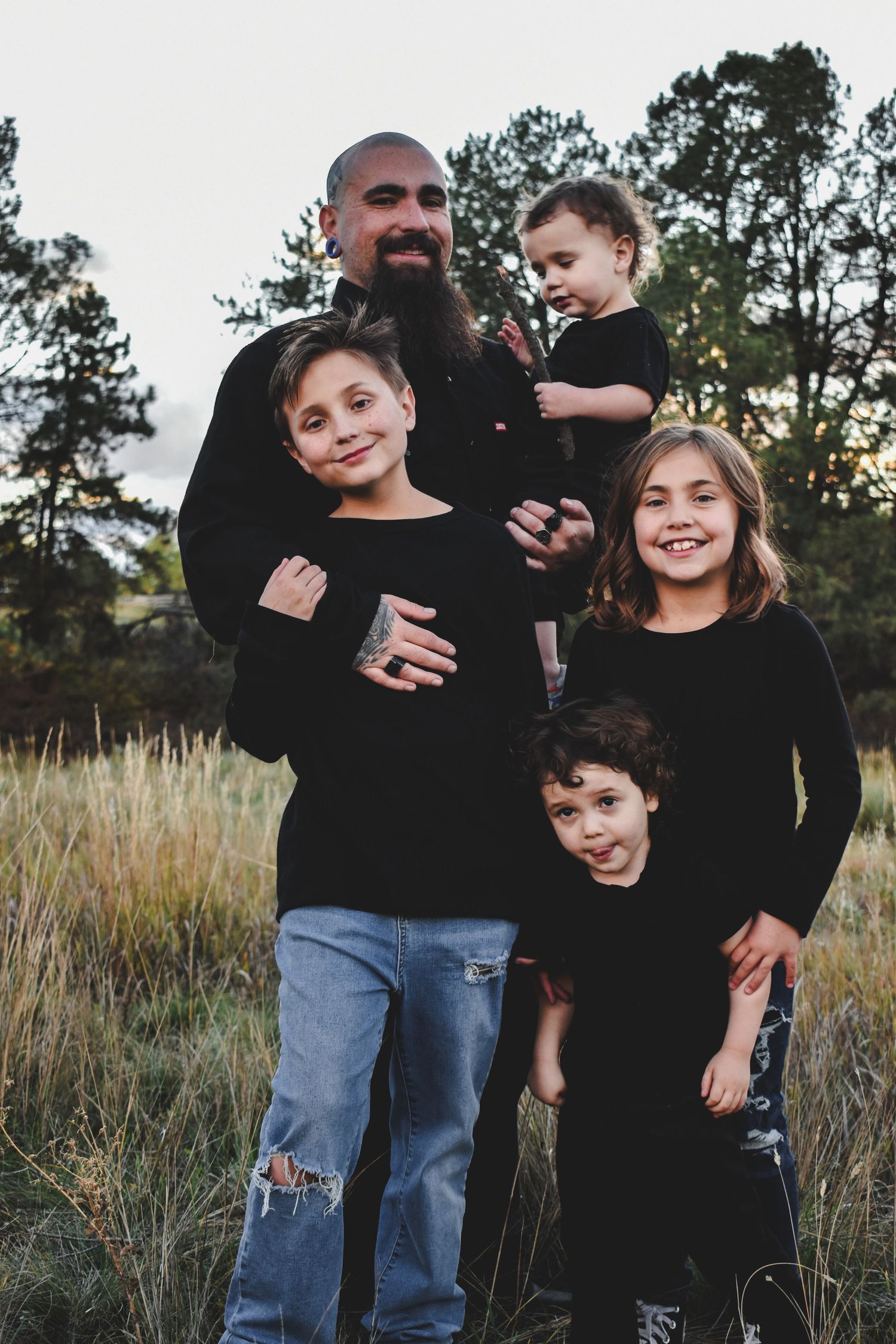 A bearded father in black poses warmly with his four children in a grassy field during golden hour sunset.