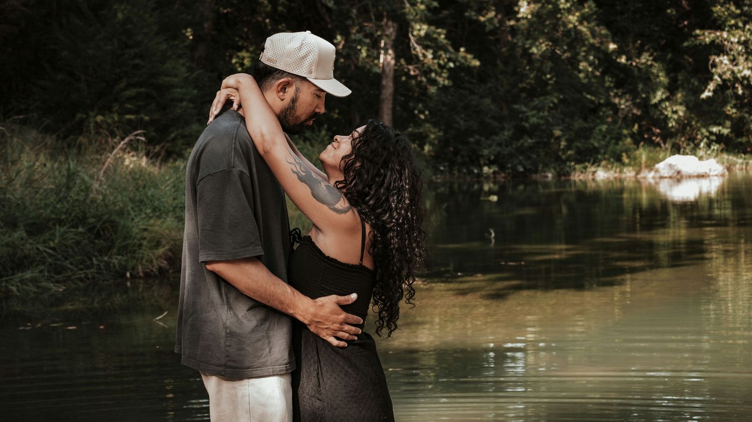 Couple sharing a romantic embrace near a serene lake surrounded by green trees in soft natural light.