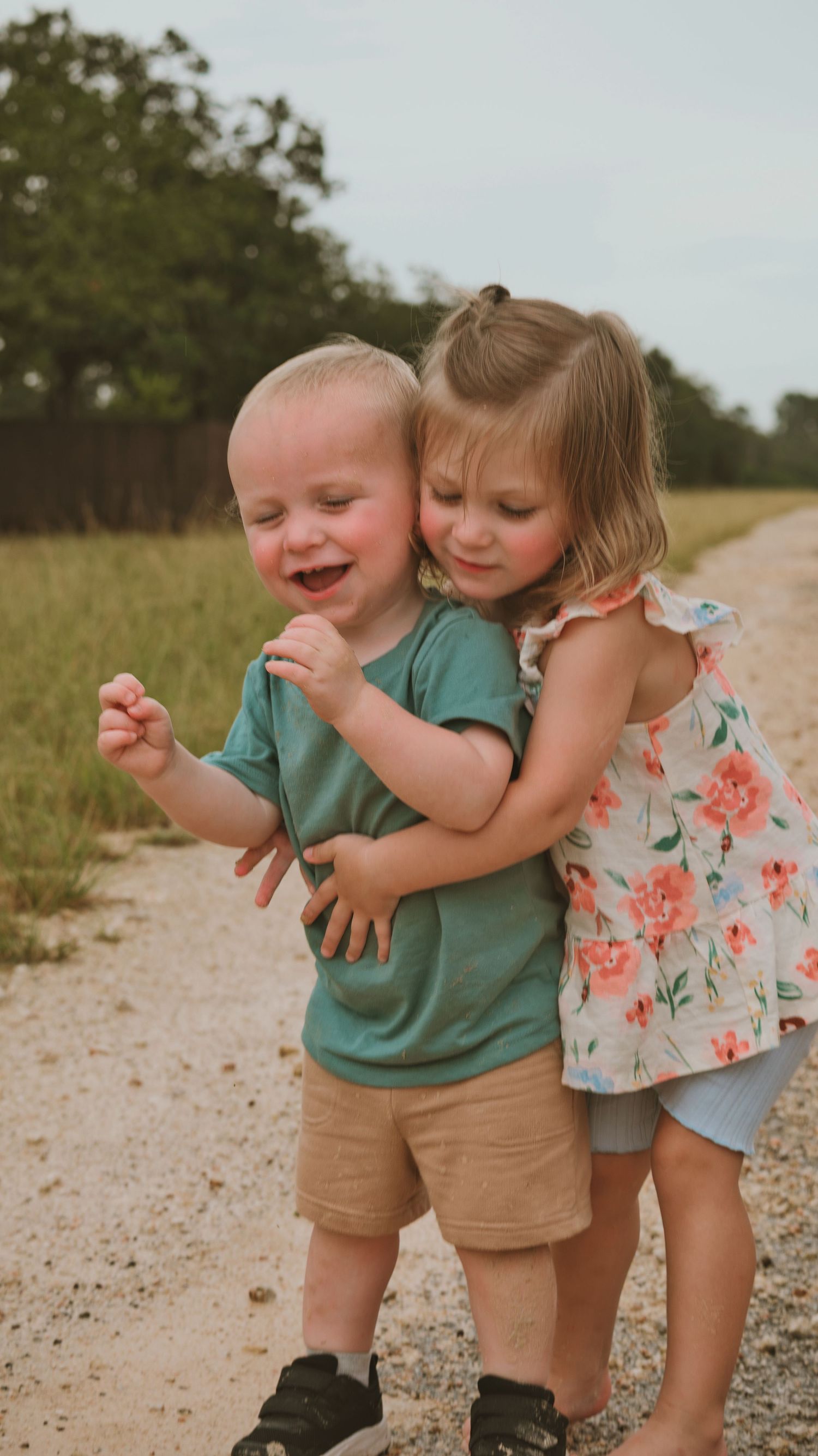 Young girl hugging her laughing baby brother from behind on a gravel country road outdoors.