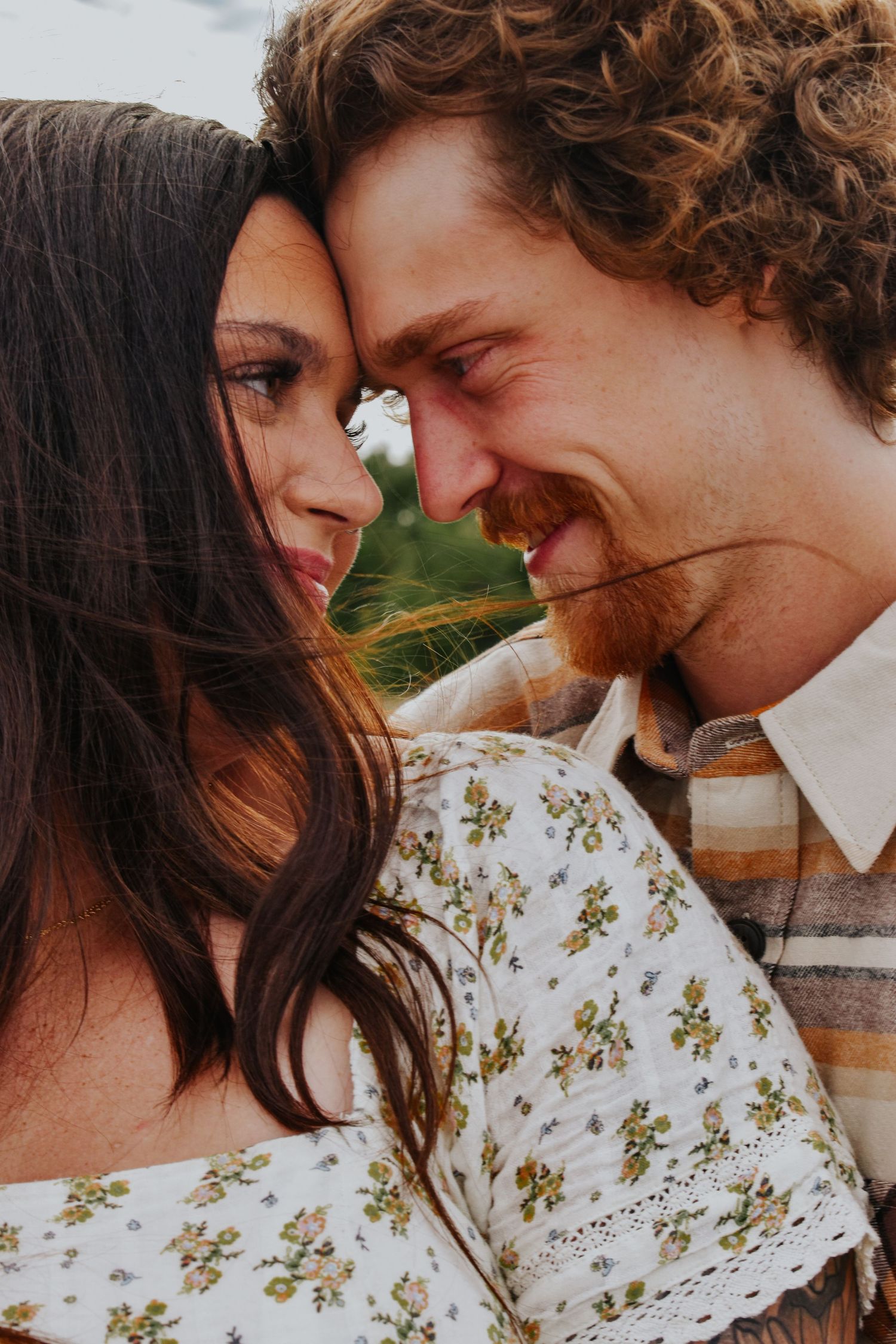 A close-up of a couple touching foreheads and smiling, the woman in a floral dress outdoors.