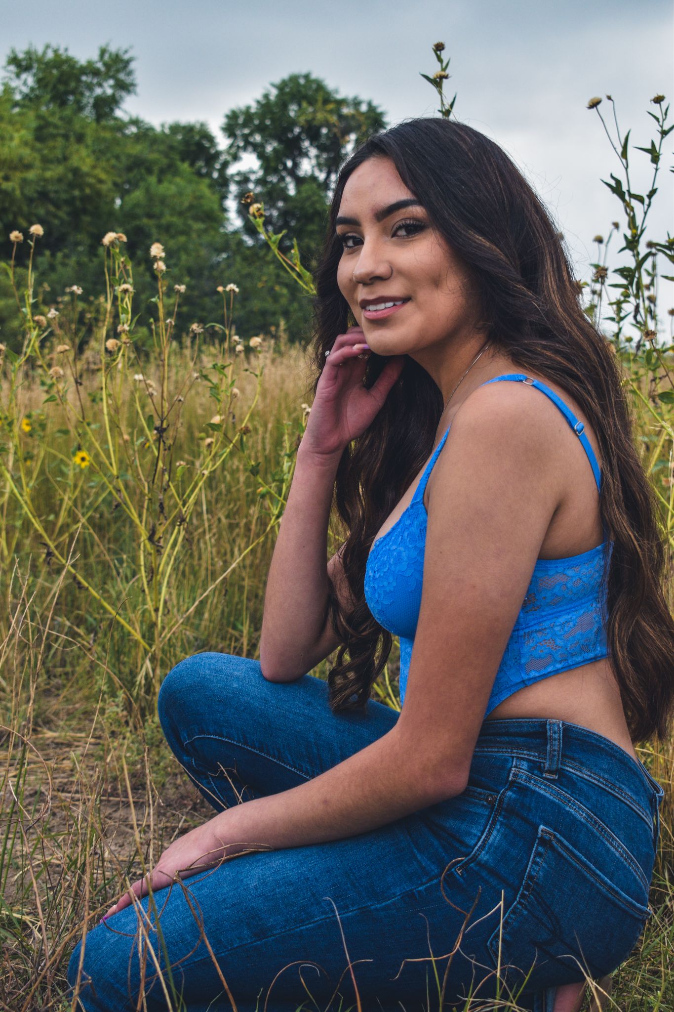Young woman in blue crop top and jeans sitting in a wildflower field, smiling at camera for senior photos.