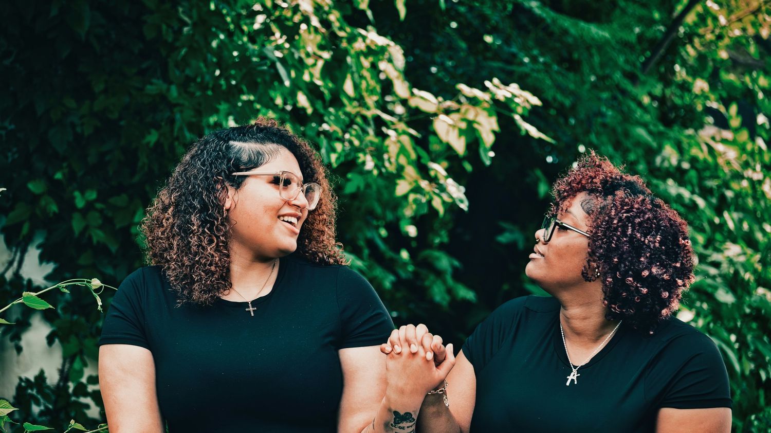 Two women in matching black outfits holding hands and smiling in a lush green setting for senior photos.