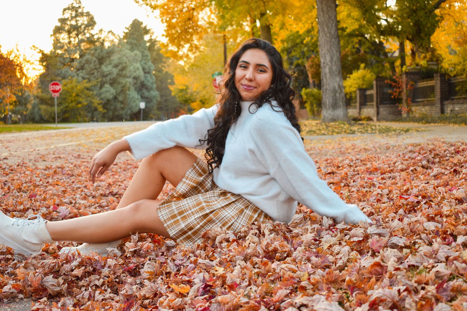Young woman in white sweater and plaid skirt sitting among colorful fallen autumn leaves in park for senior photos.