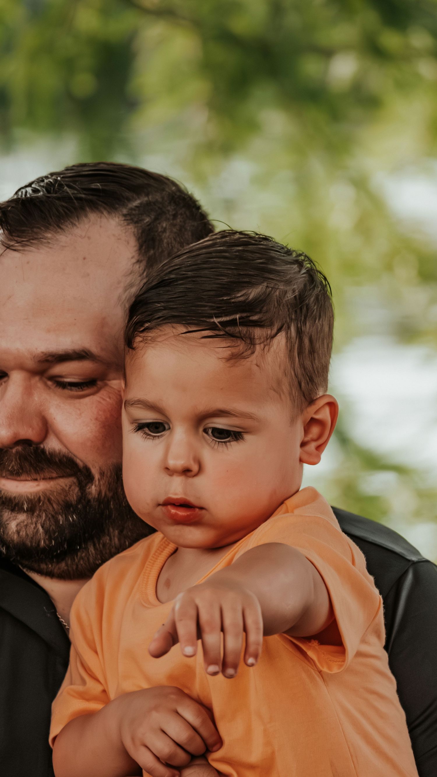 A bearded father tenderly holds his son outdoors with green trees behind.