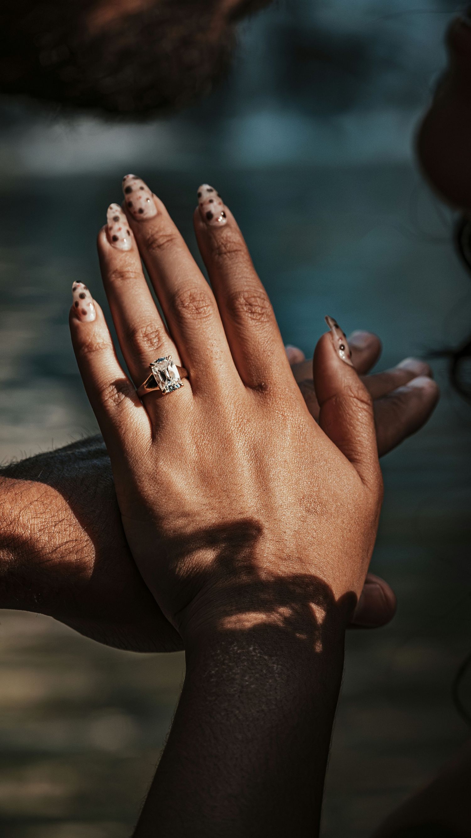 Close-up of a woman's hand with spotted nail art and a ring held by her partner near water for a couples session.