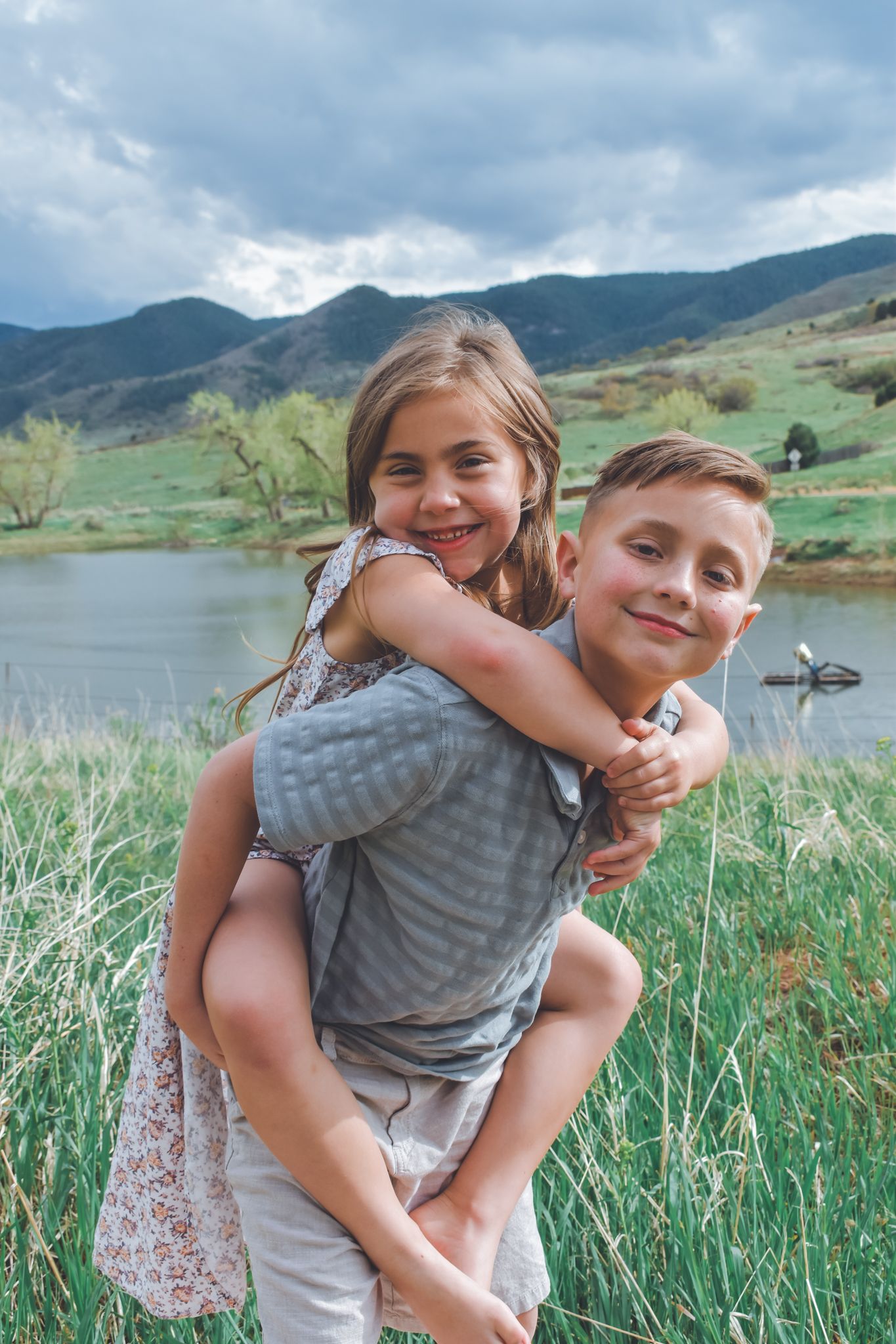 Smiling girl rides piggyback on a boy in front of a mountain lake and green hills. Family Photographer in Pflugerville, Tx