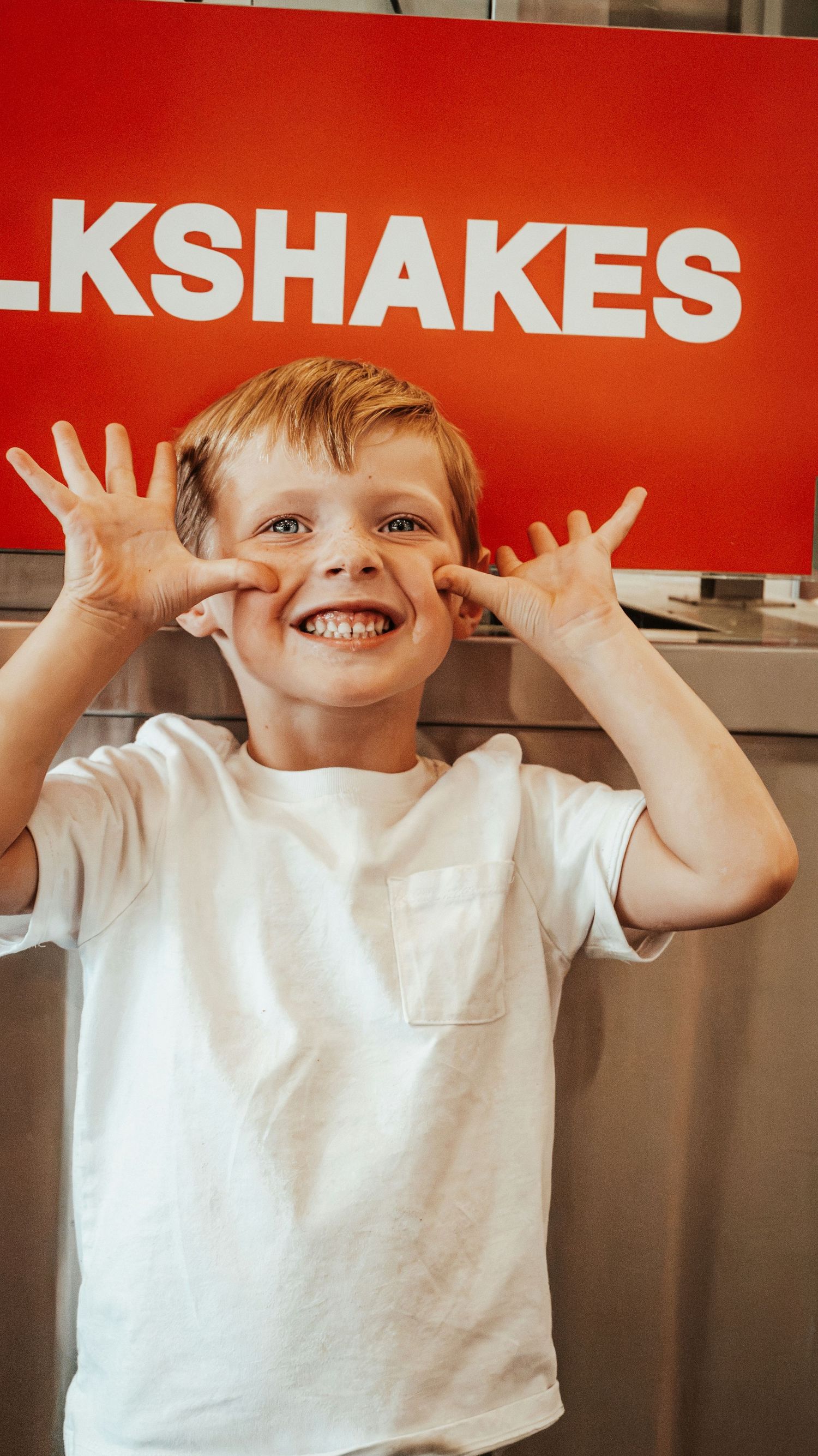 A smiling blond boy makes a funny face in front of a red milkshakes sign at Five Guys restaurant. Family Photographer in Pflugerville, Tx