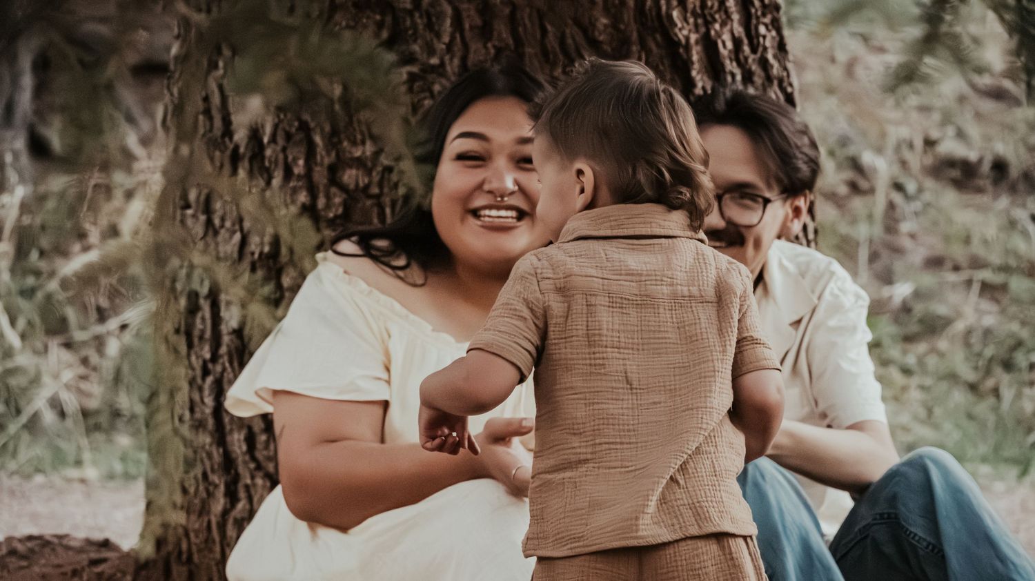 A joyful mother laughs as her toddler reaches for her while the family sits by a large tree.