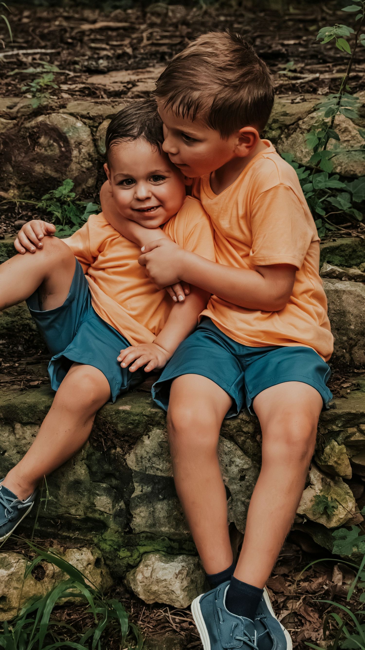Older boy in orange shirt hugs and kisses younger sibling while sitting together on a rustic stone wall.
