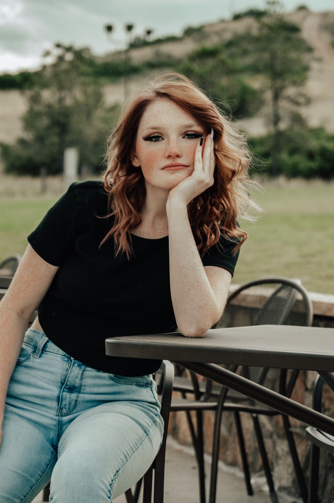 Redhead woman with freckles in black tee and light jeans resting chin on hand at outdoor patio table for senior photos.