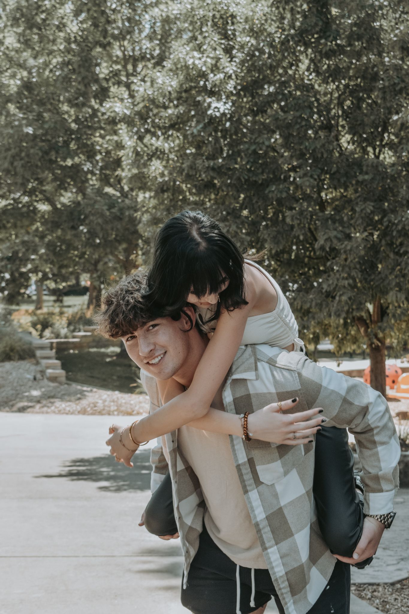Smiling young man gives a laughing dark-haired woman a piggyback ride on a sunny outdoor pathway for senior photos.