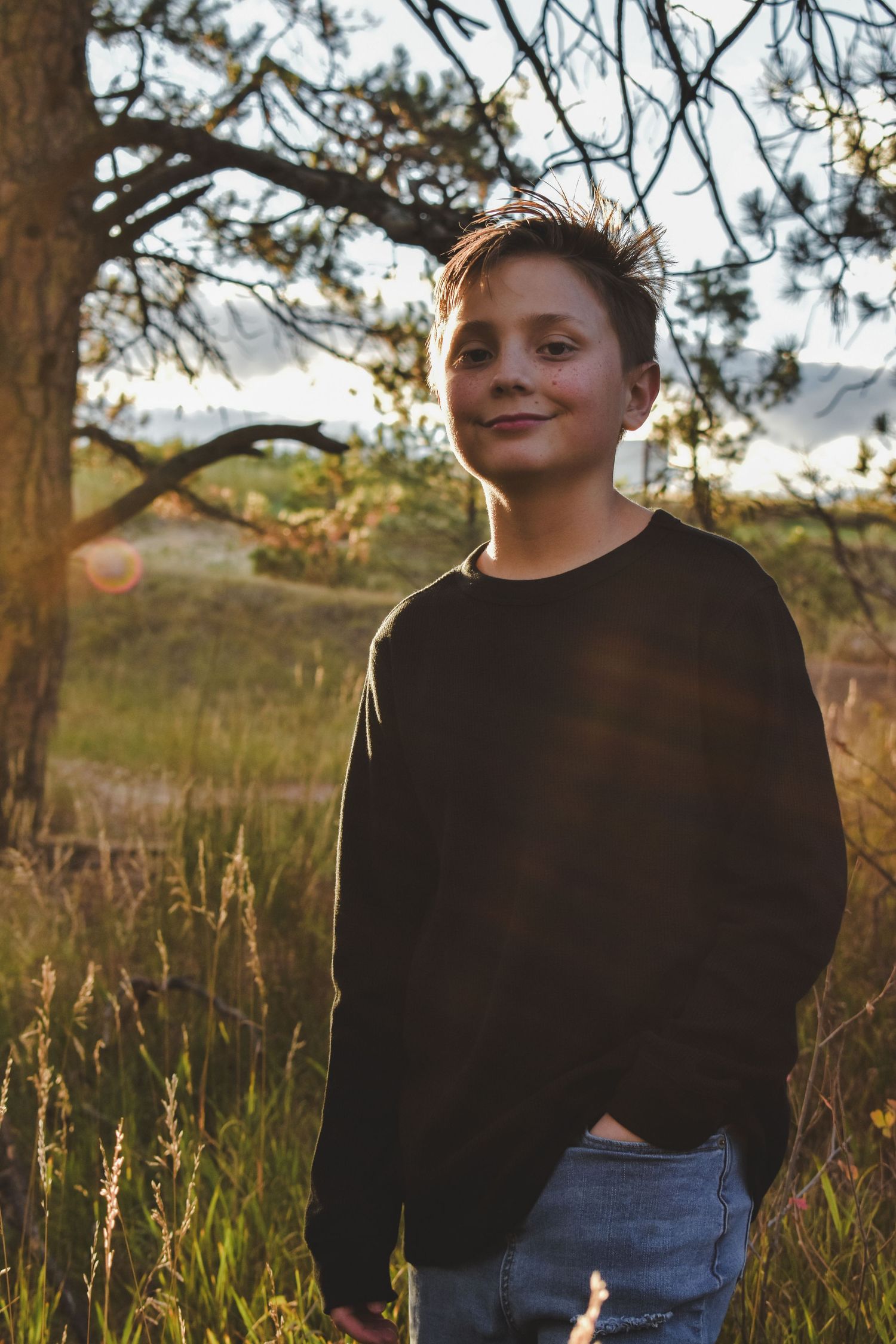 Smiling preteen boy in black sweater standing in golden sunlit field near pine trees.