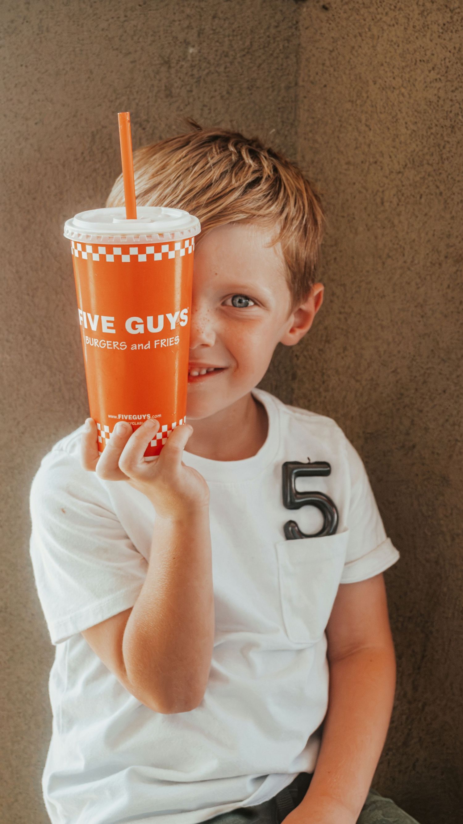 Smiling boy in white t-shirt holding an orange Five Guys drink cup with straw.