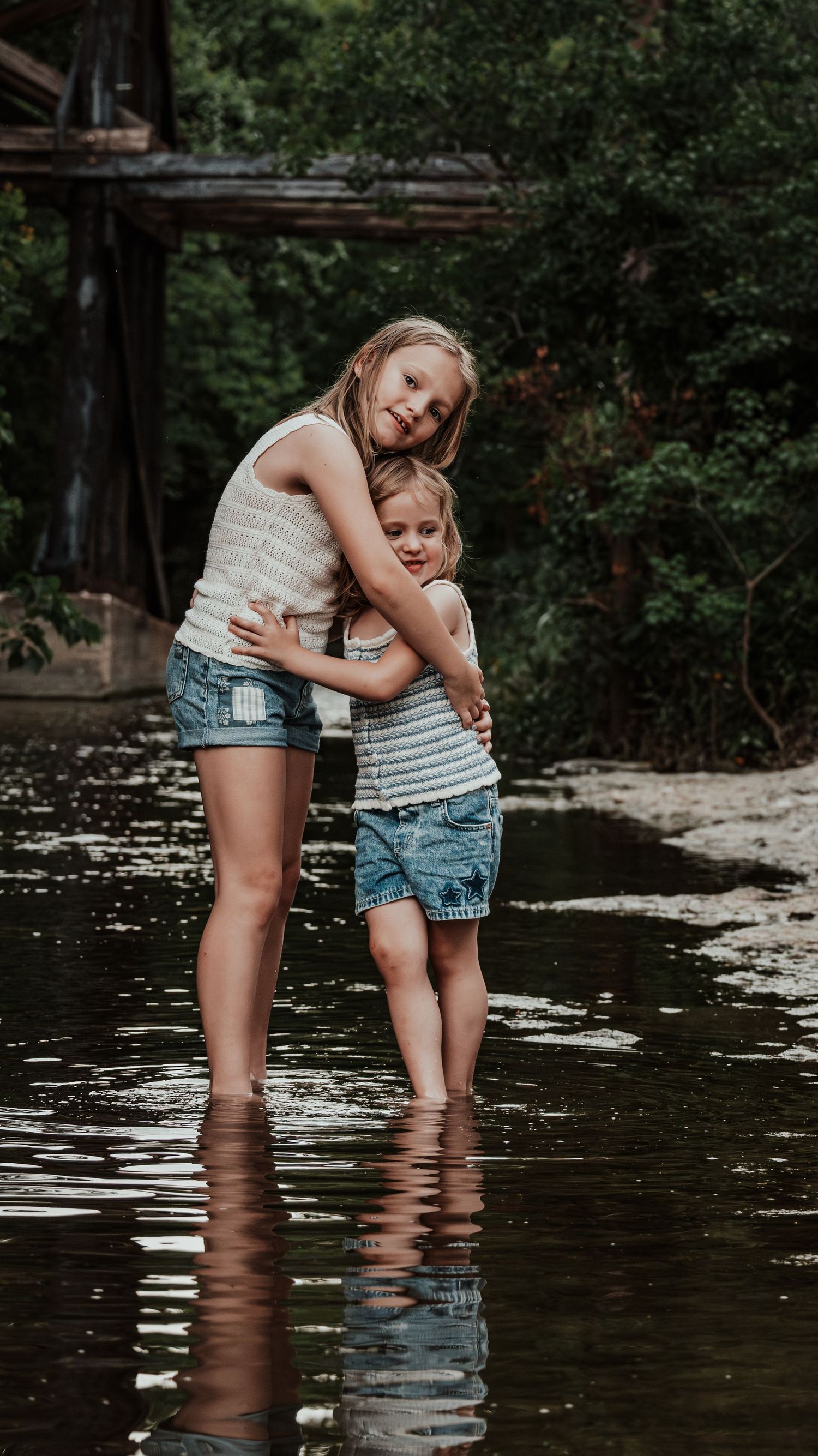 Two girls in matching tank tops and denim shorts share a warm hug while standing in a creek.
