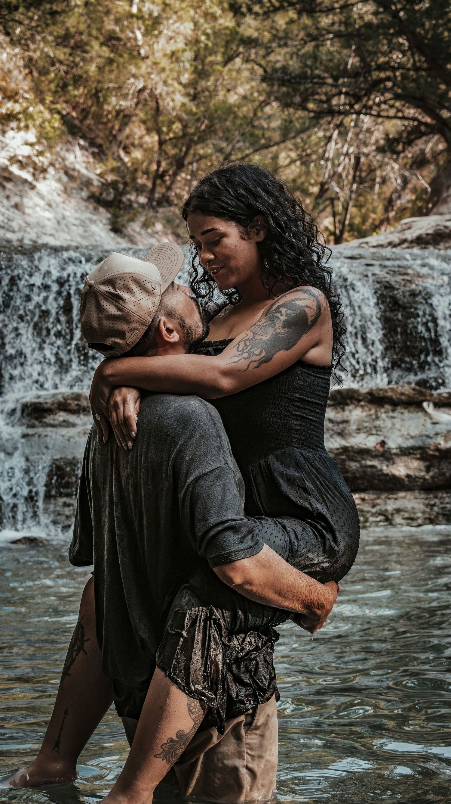 Man carrying woman in his arms while she smiles, standing in water with waterfall behind them.
