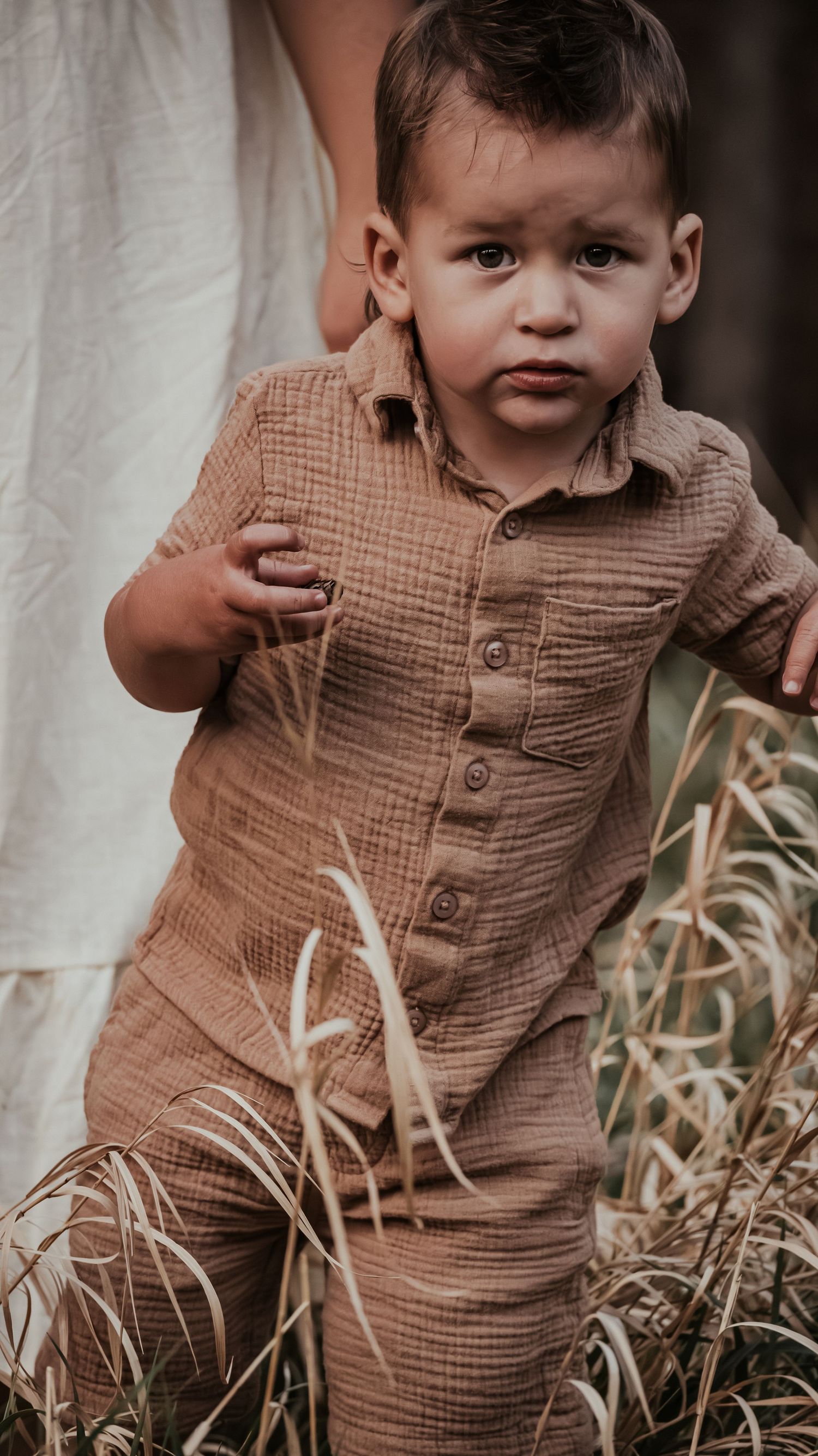 Young toddler boy in a brown linen matching outfit walking through tall grass near his mother.