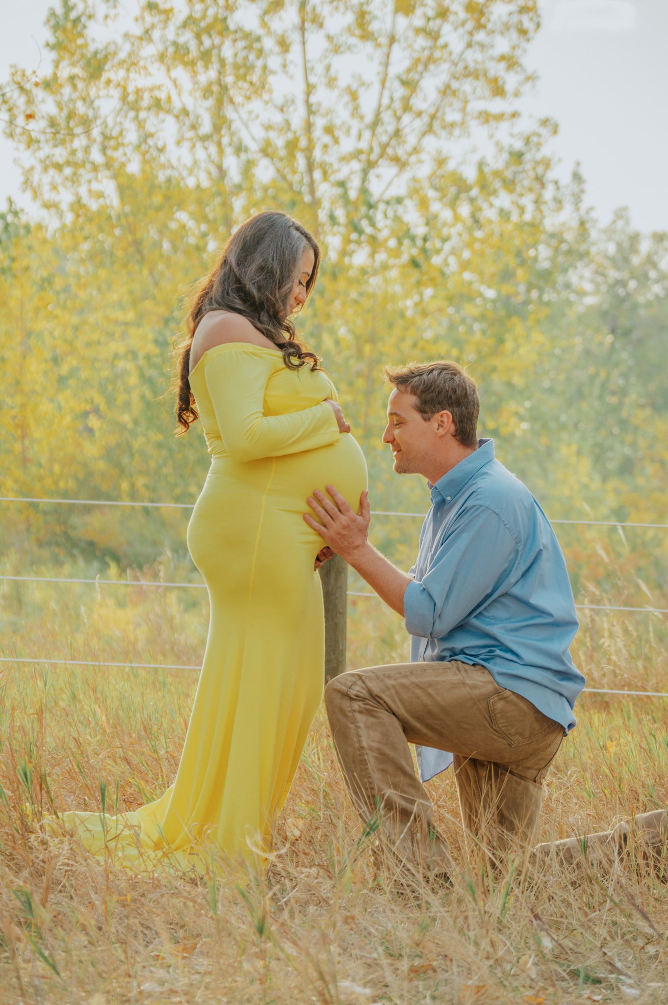 Man kneeling to touch pregnant womans belly in yellow dress in field.