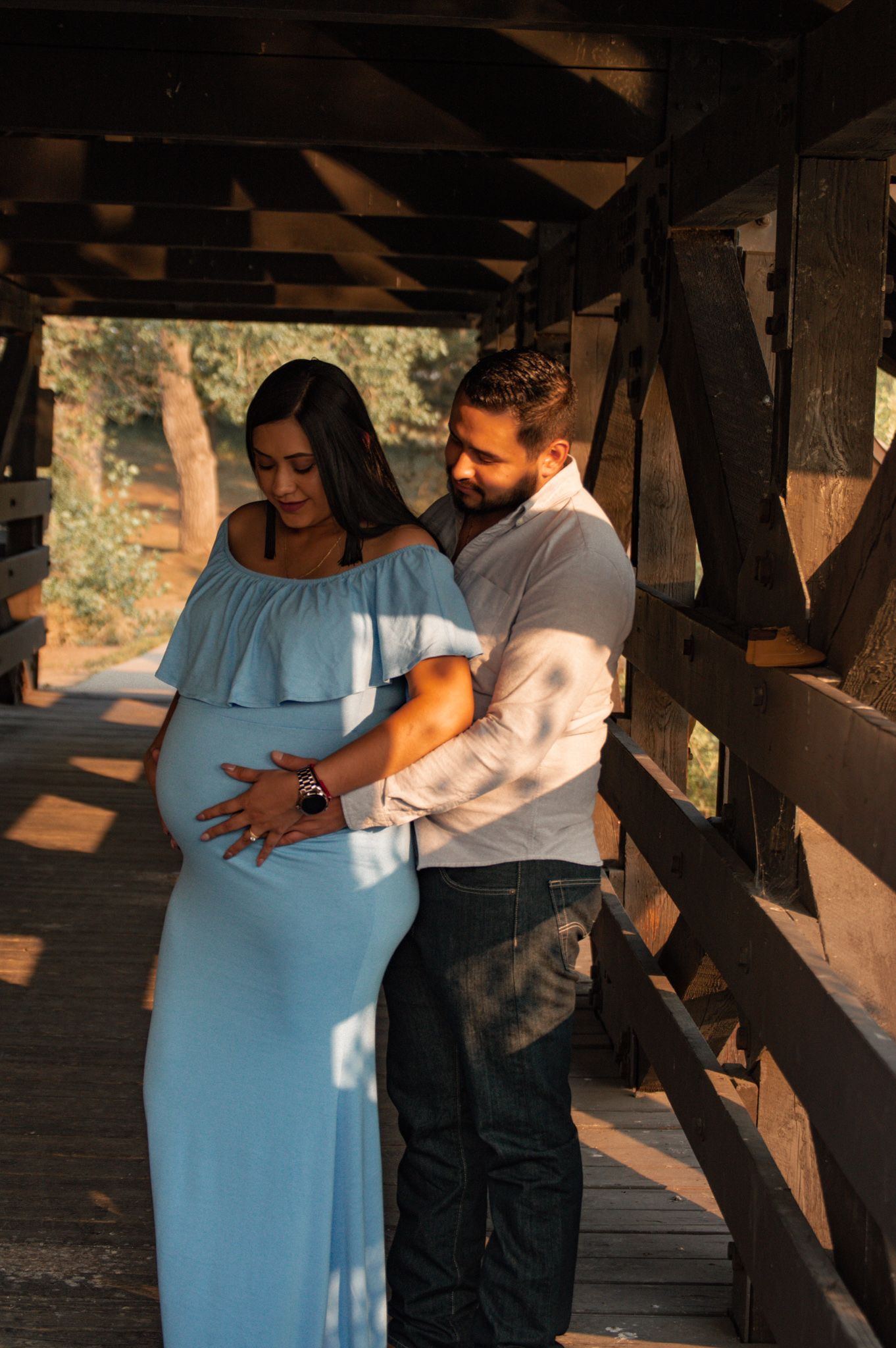 Couple embracing on wooden bridge at golden hour, woman in blue off-shoulder maternity gown.