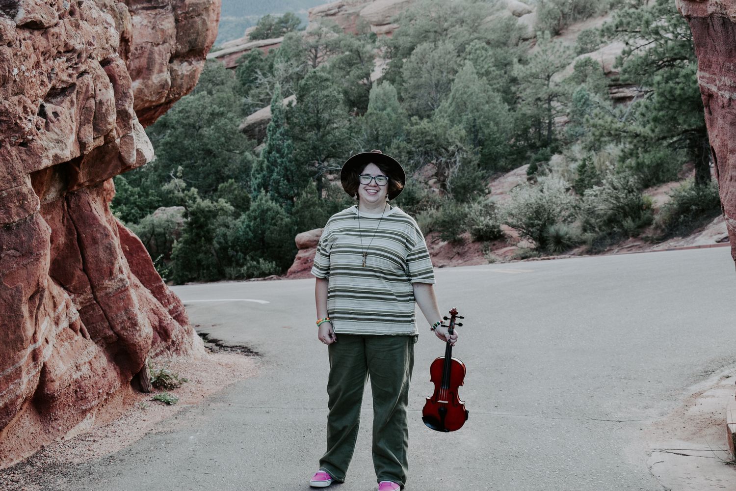 Person standing near red rocks holding a violin, wearing a striped shirt, hat, and pink shoes for senior photos.
