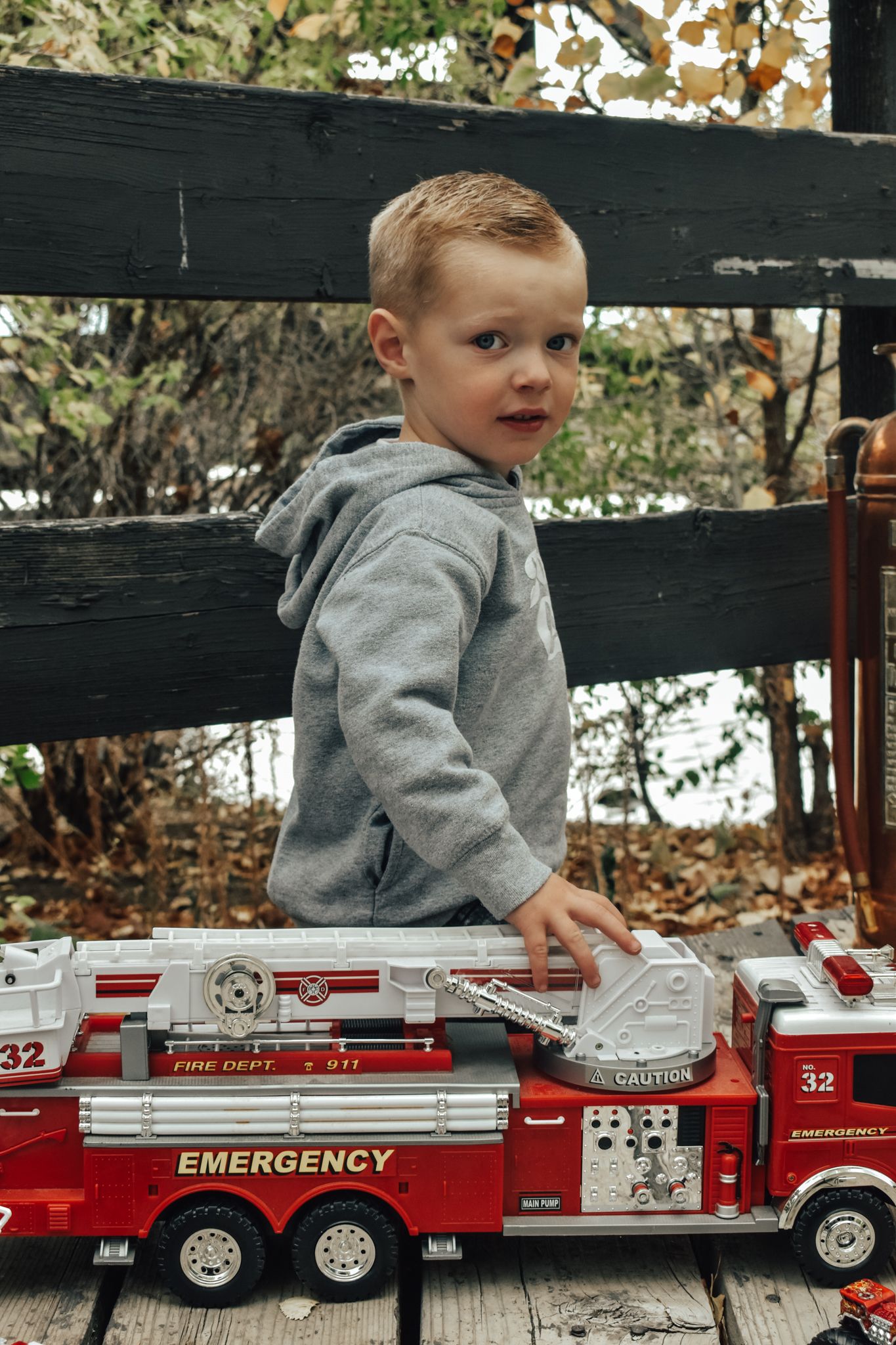 Young boy in gray hoodie plays with red toy emergency fire truck on wooden deck outdoors. Family Photographer in Pflugerville, Tx