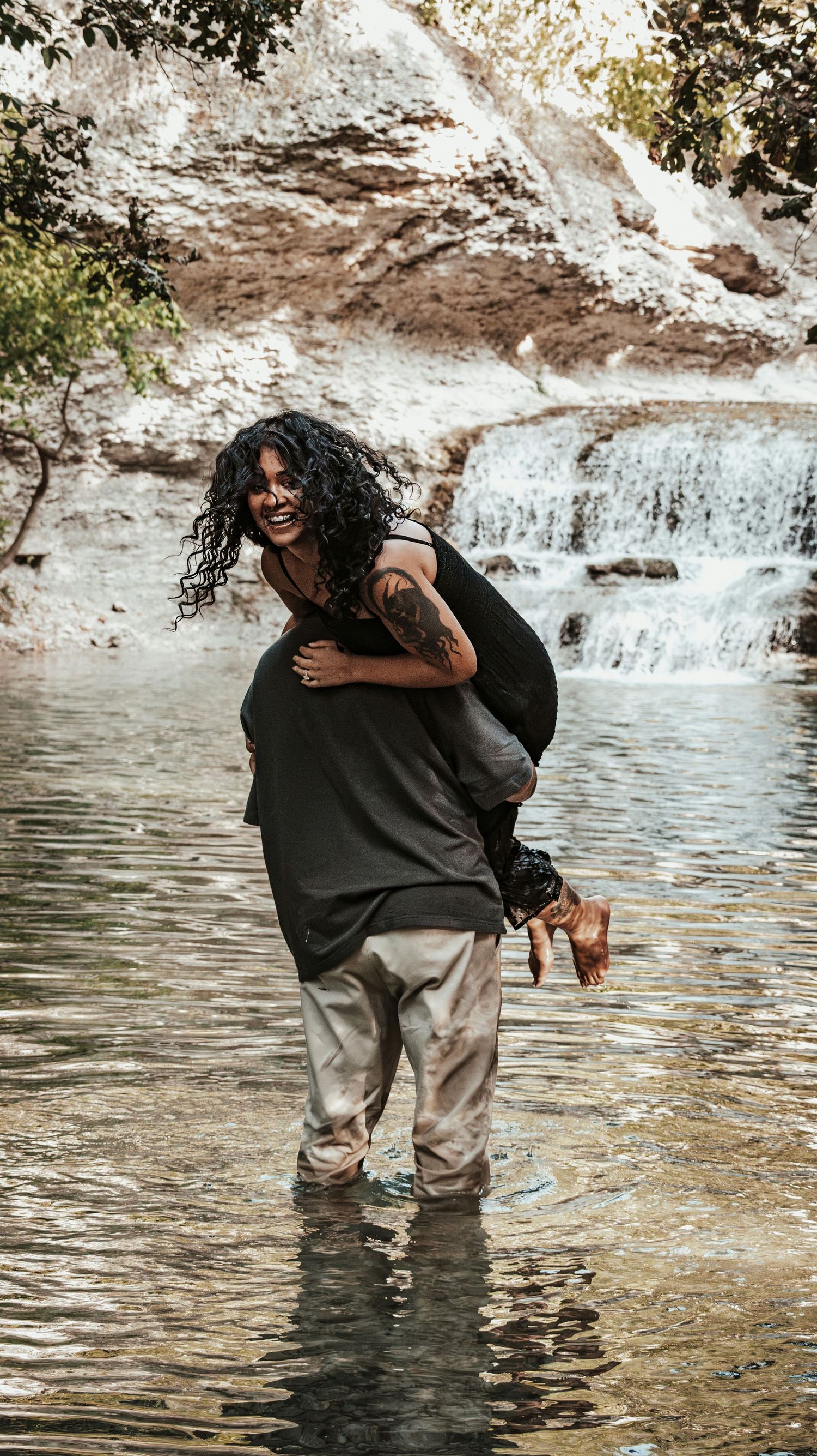 A man carrying a laughing woman on his back while wading through a shallow river near a waterfall for a couples session.