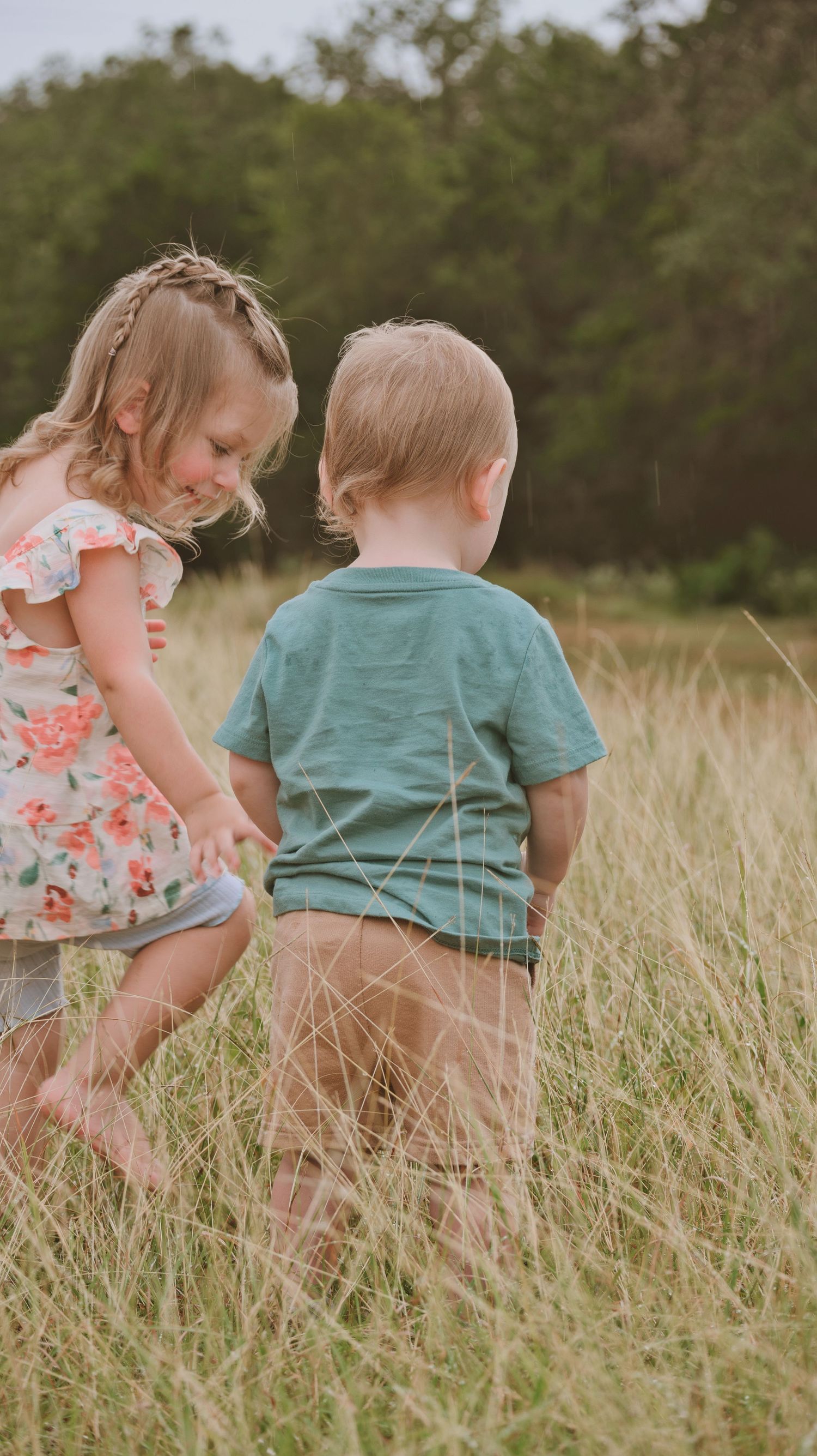Two young blonde children playing together in a tall grass field with trees in the background for a family session.