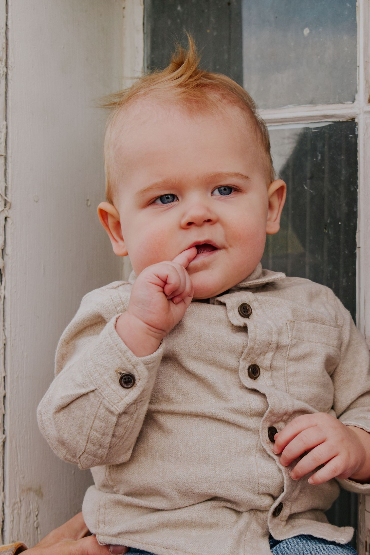 Adorable blue-eyed baby in linen shirt sitting by an old window frame.