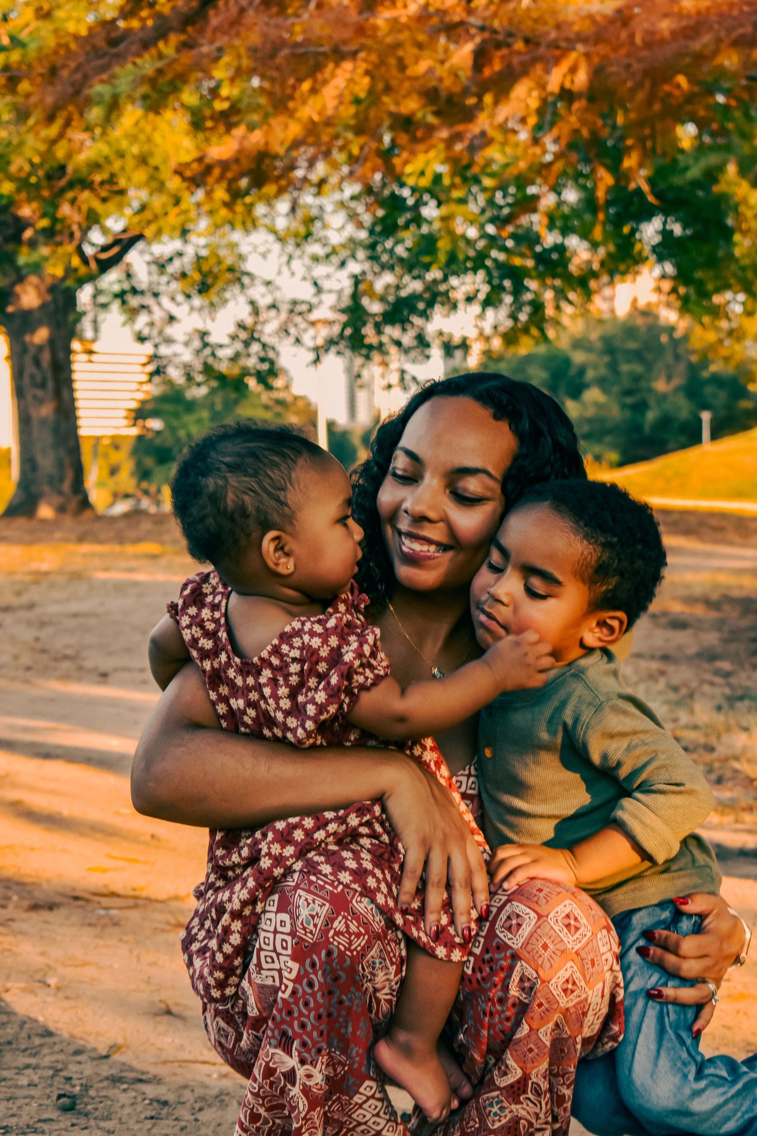 A smiling mother holds her two young children in a sunlit autumn park, sharing a warm family embrace.