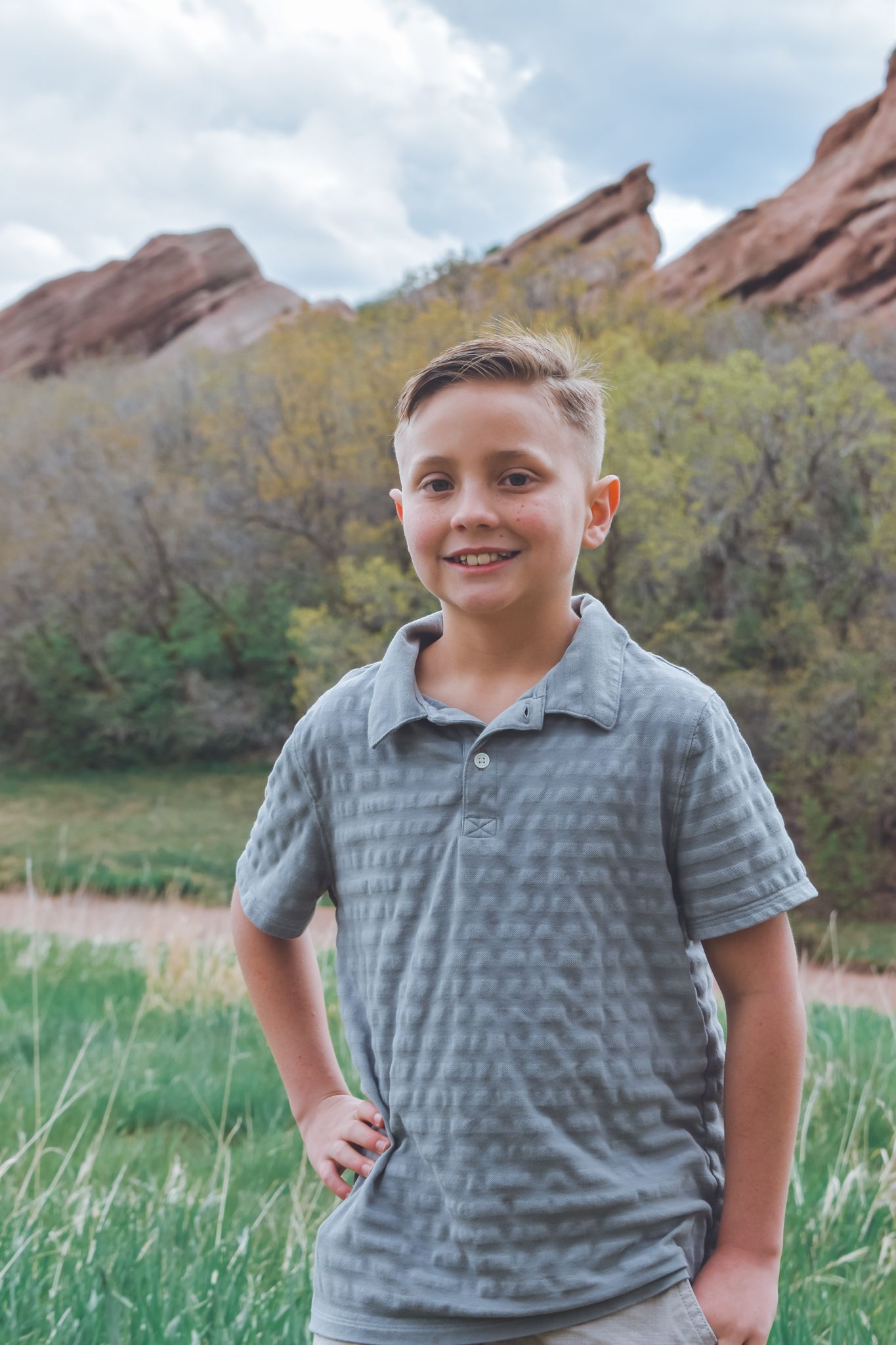 Smiling boy in gray polo shirt standing in front of red rock formations at Red Rocks Park, Colorado.
