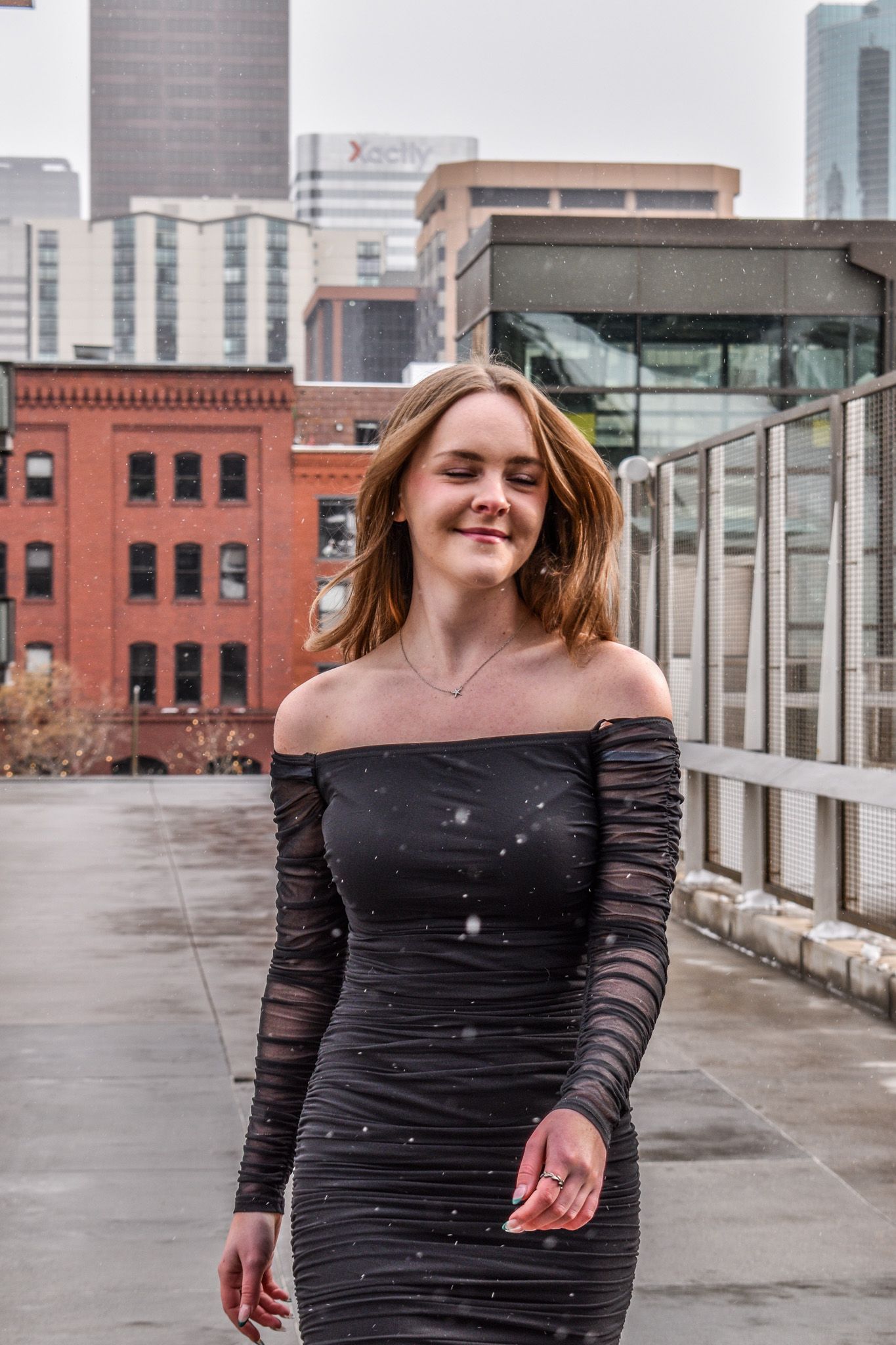 Smiling young woman in black off-shoulder dress posing on rooftop with city buildings backdrop for senior photos for senior photos.