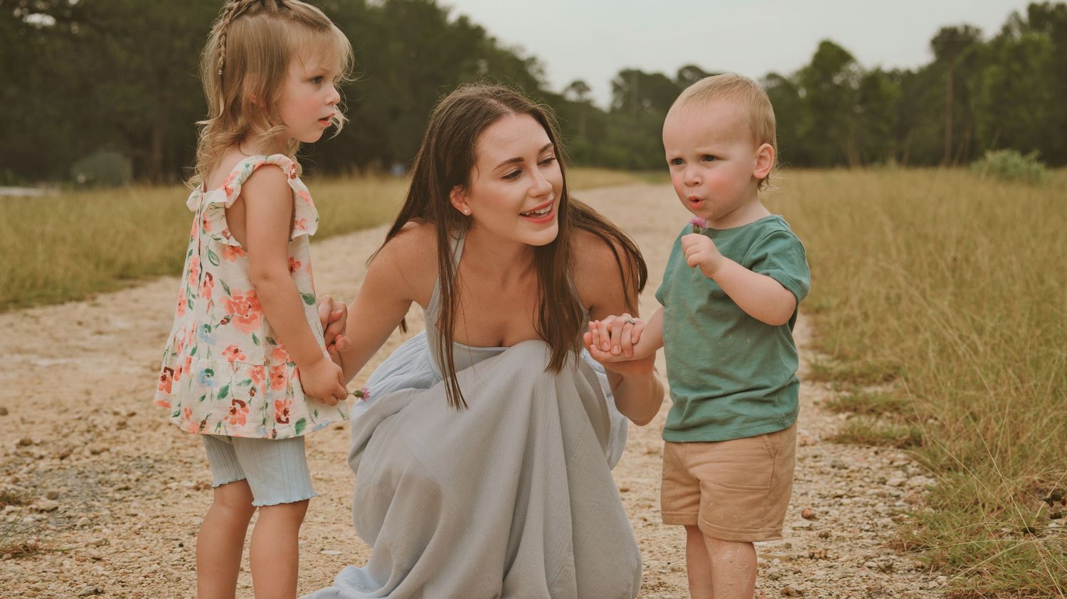 Mother kneels on gravel path holding hands with toddler daughter and young son outdoors.