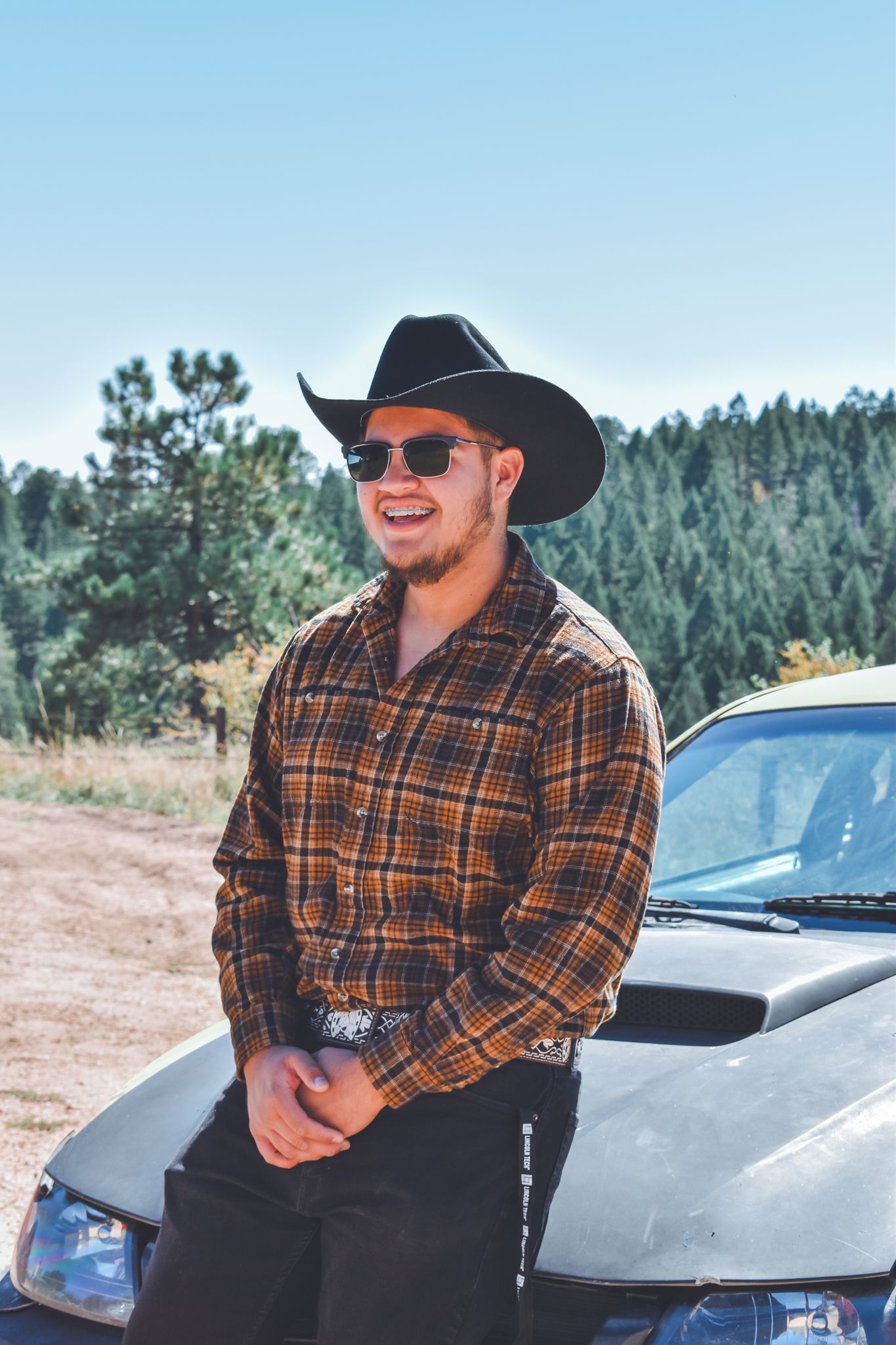 Smiling young man in cowboy hat and plaid shirt leaning on a dark car with pine trees behind for senior photos.