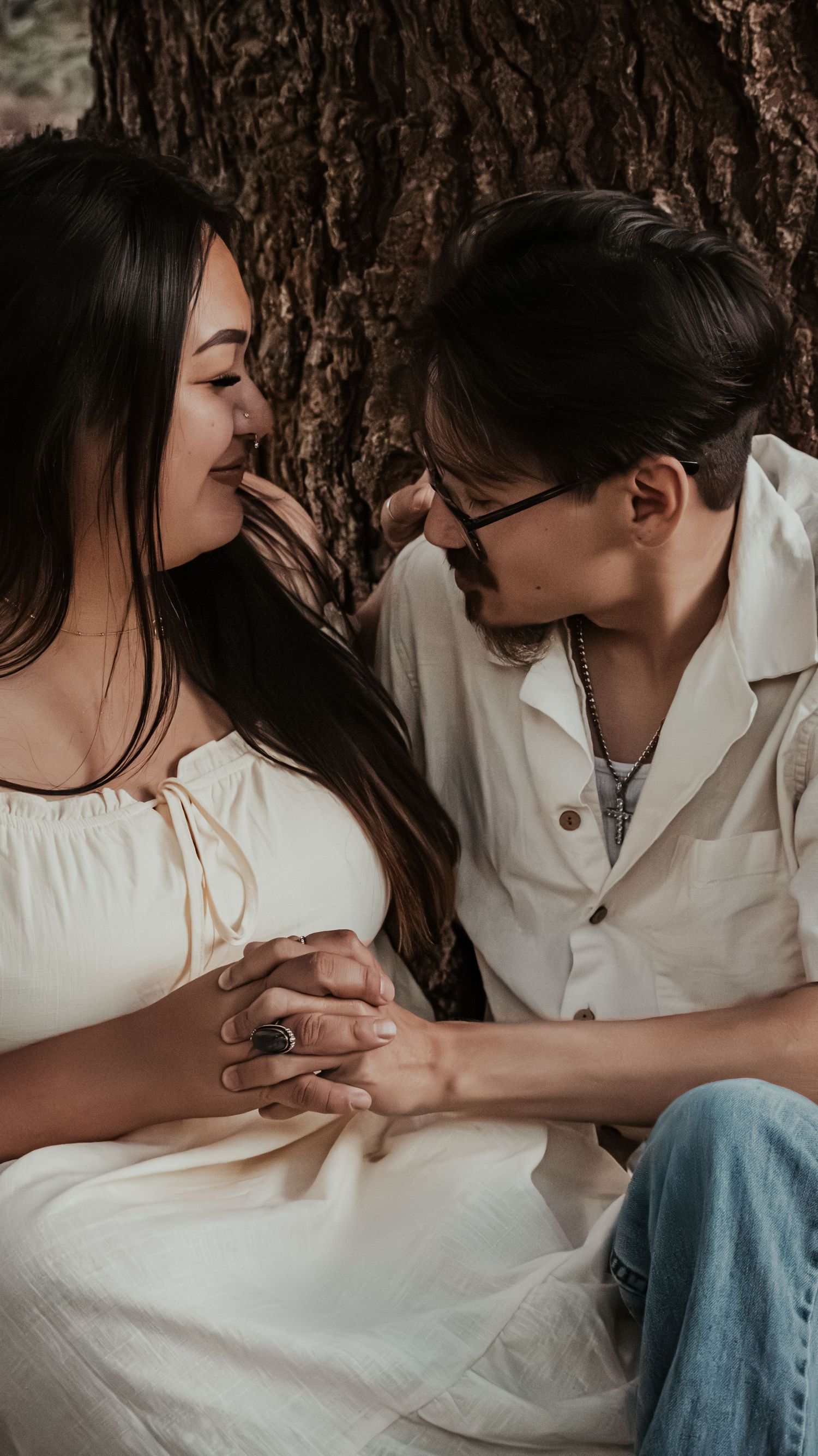 A couple holds hands and shares an intimate moment leaning against a tree trunk.