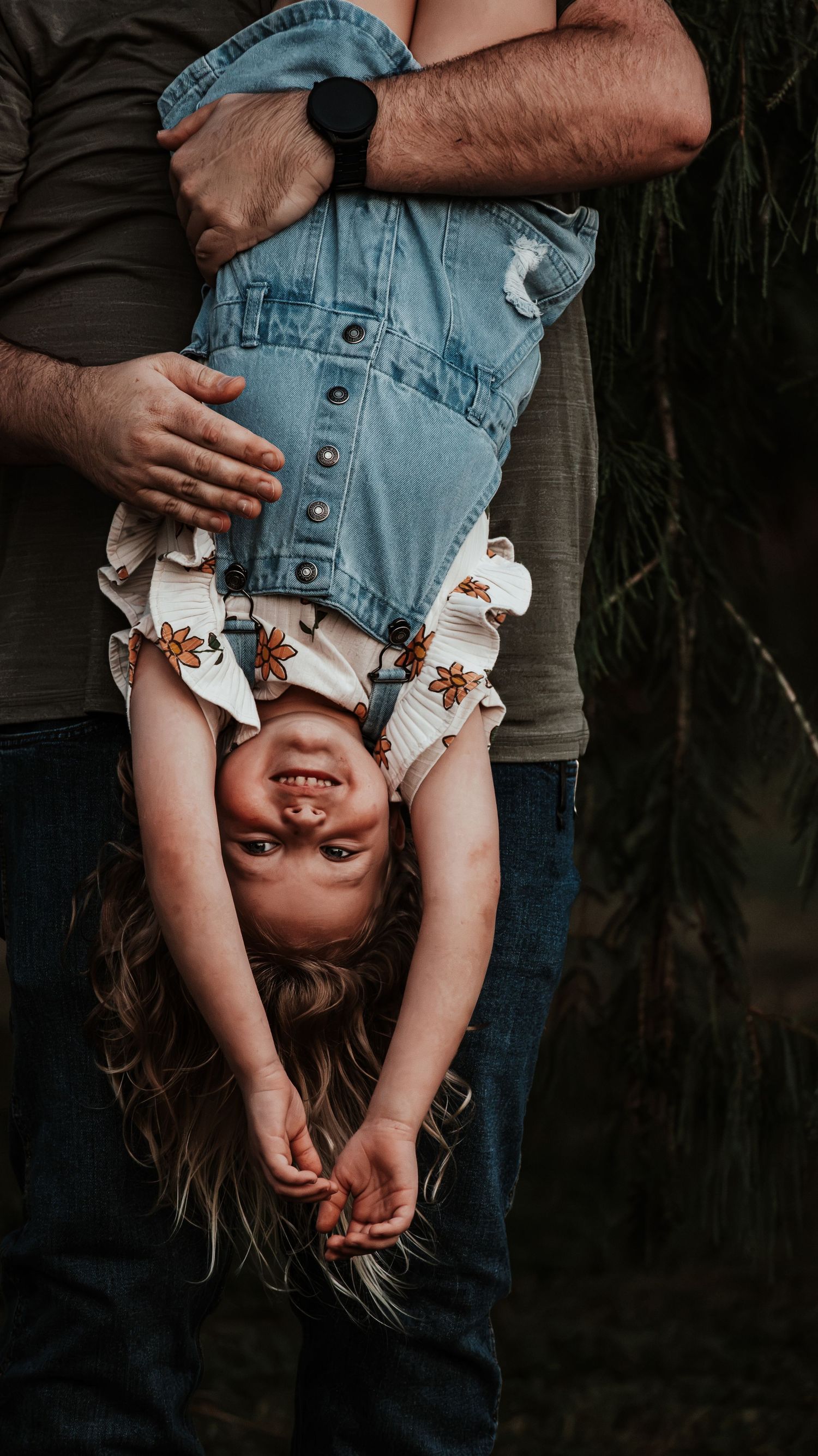 Playful toddler girl in denim overalls held upside down by parent, laughing joyfully outdoors.