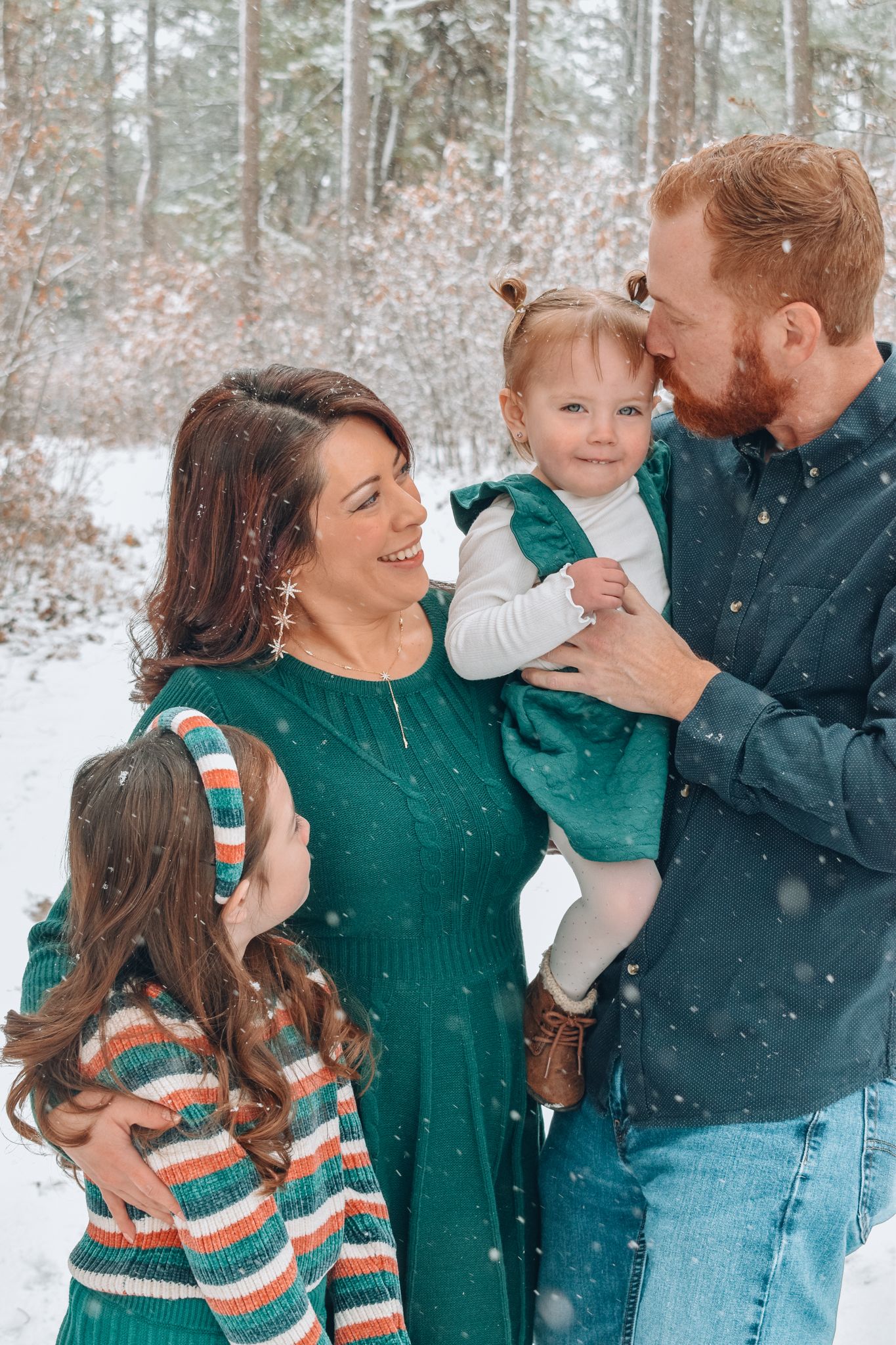 Happy family of four posing in snowy forest, parents in green outfits holding two young daughters.