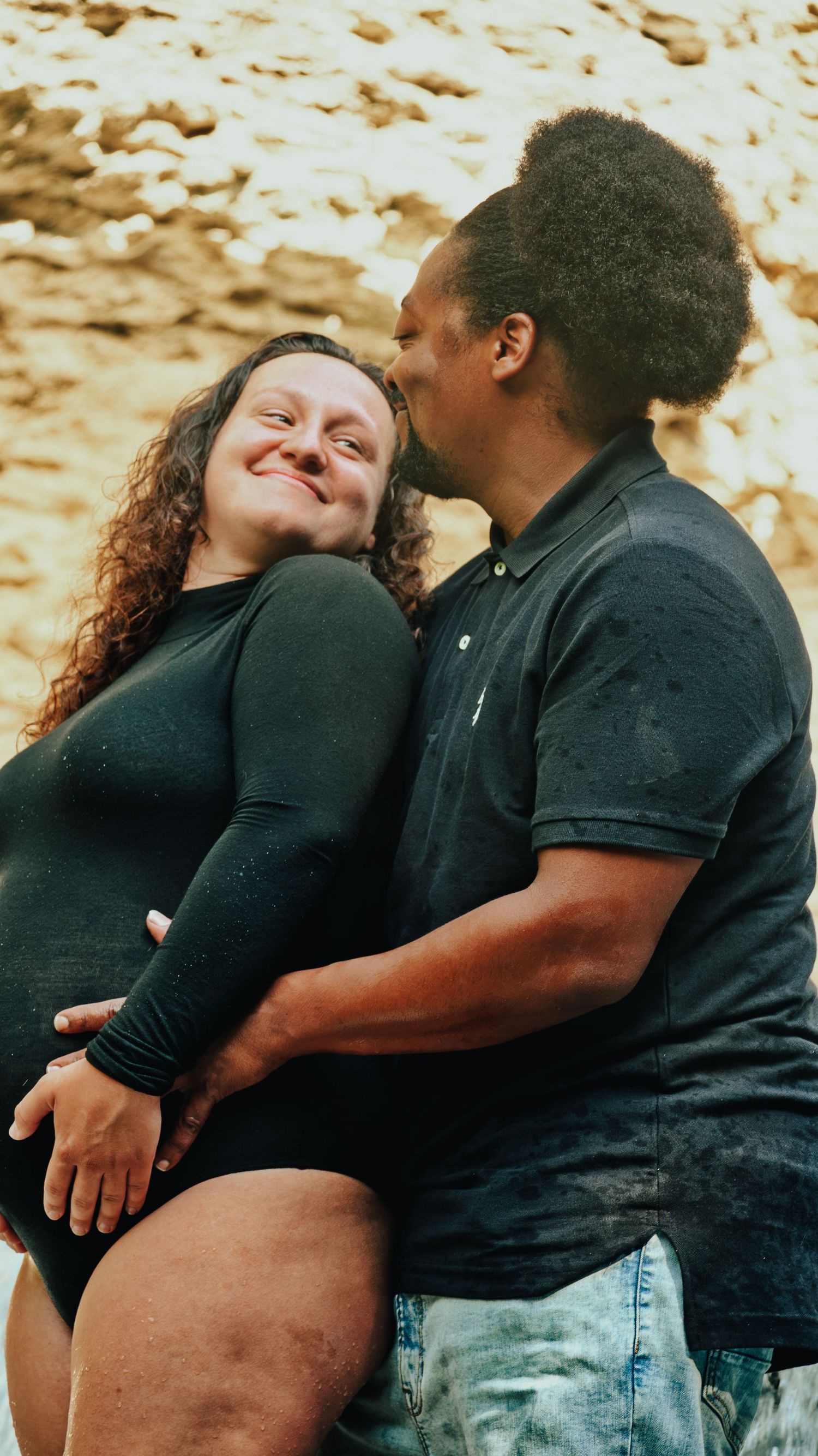 Joyful expecting couple sharing a kiss near rocky terrain, woman in black maternity bodysuit.