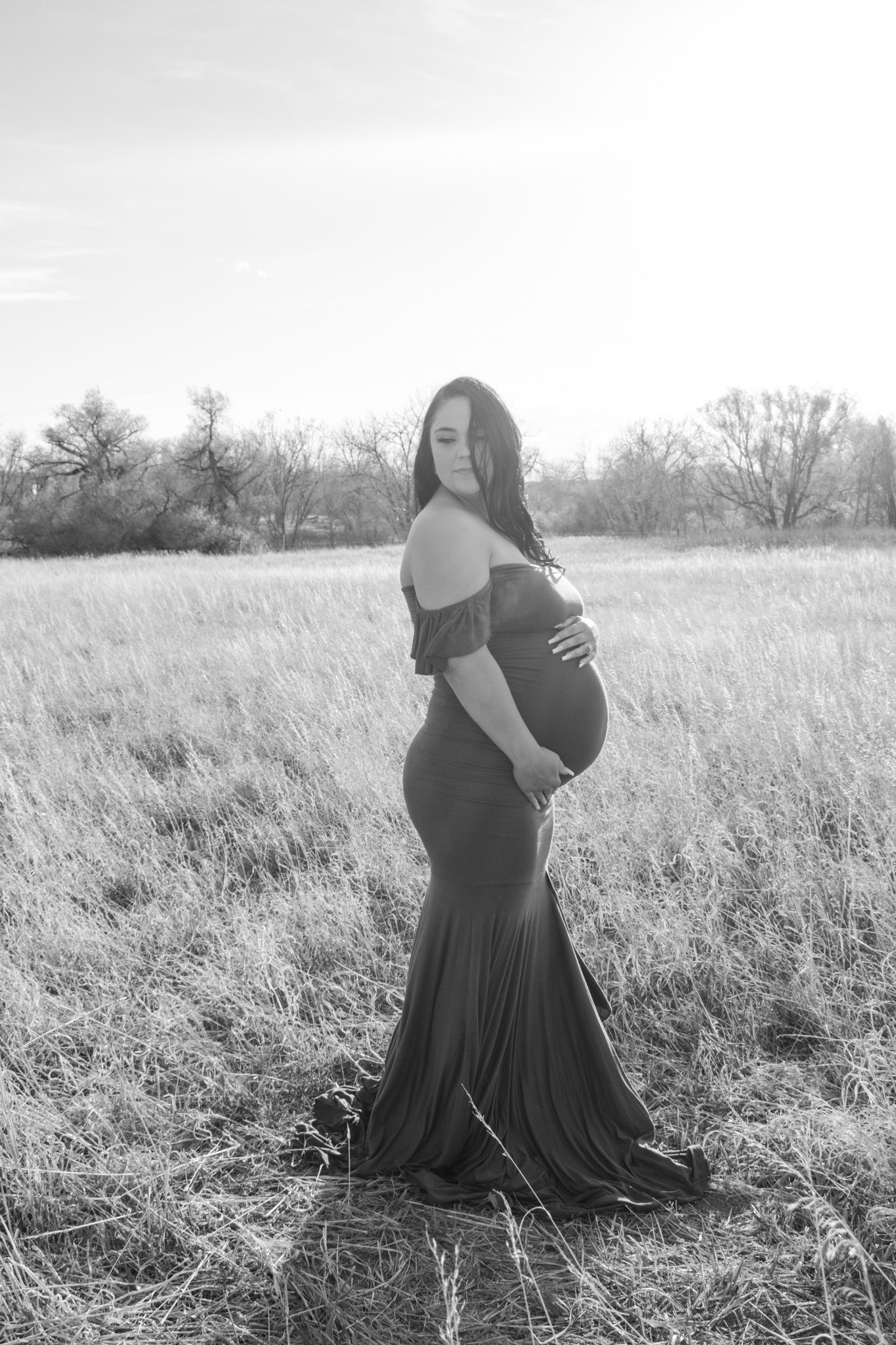 Black and white maternity portrait of tattooed pregnant woman in form-fitting gown in field.