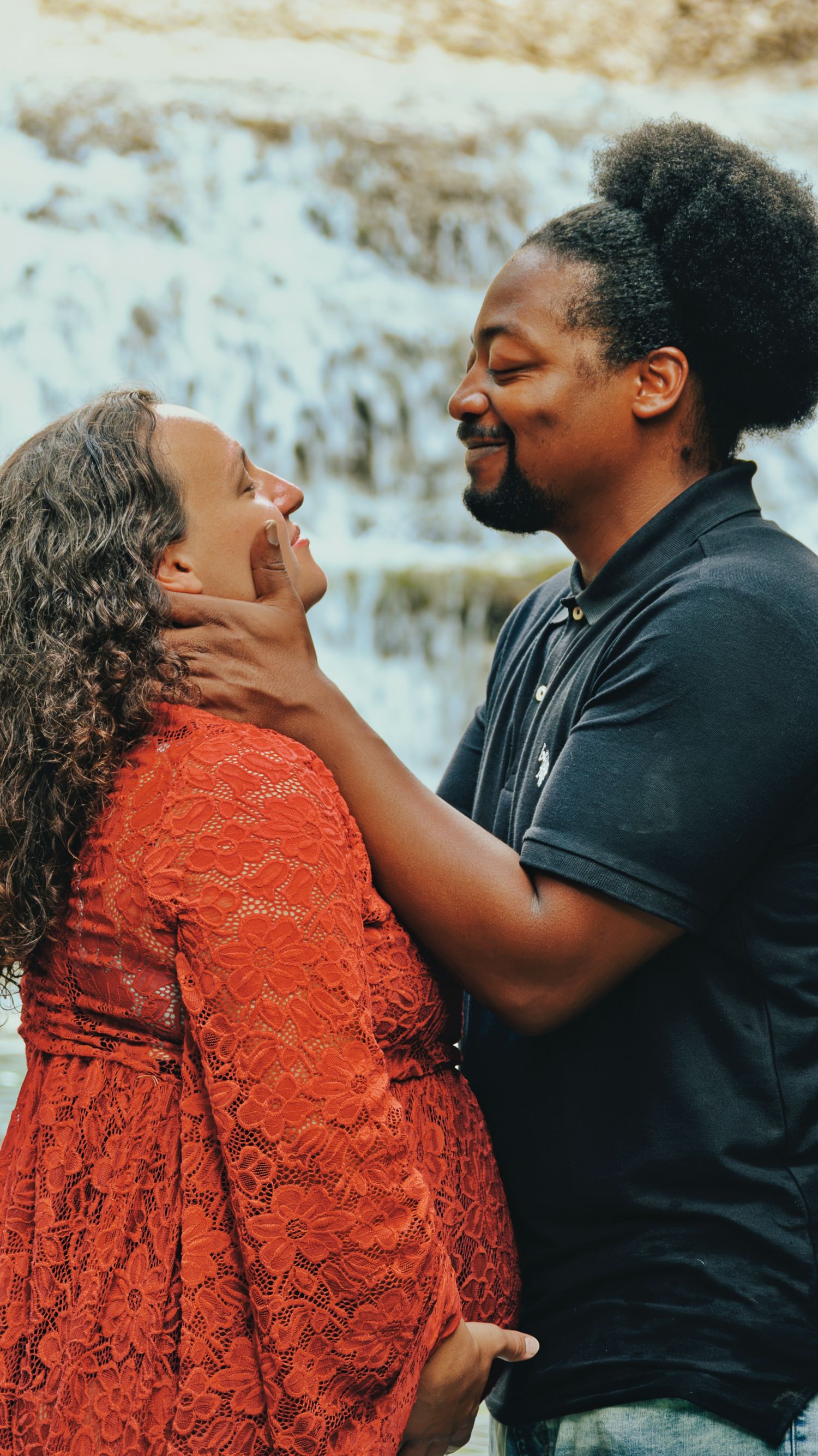 Joyful interracial couple laughs together during outdoor maternity photo session near waterfall.