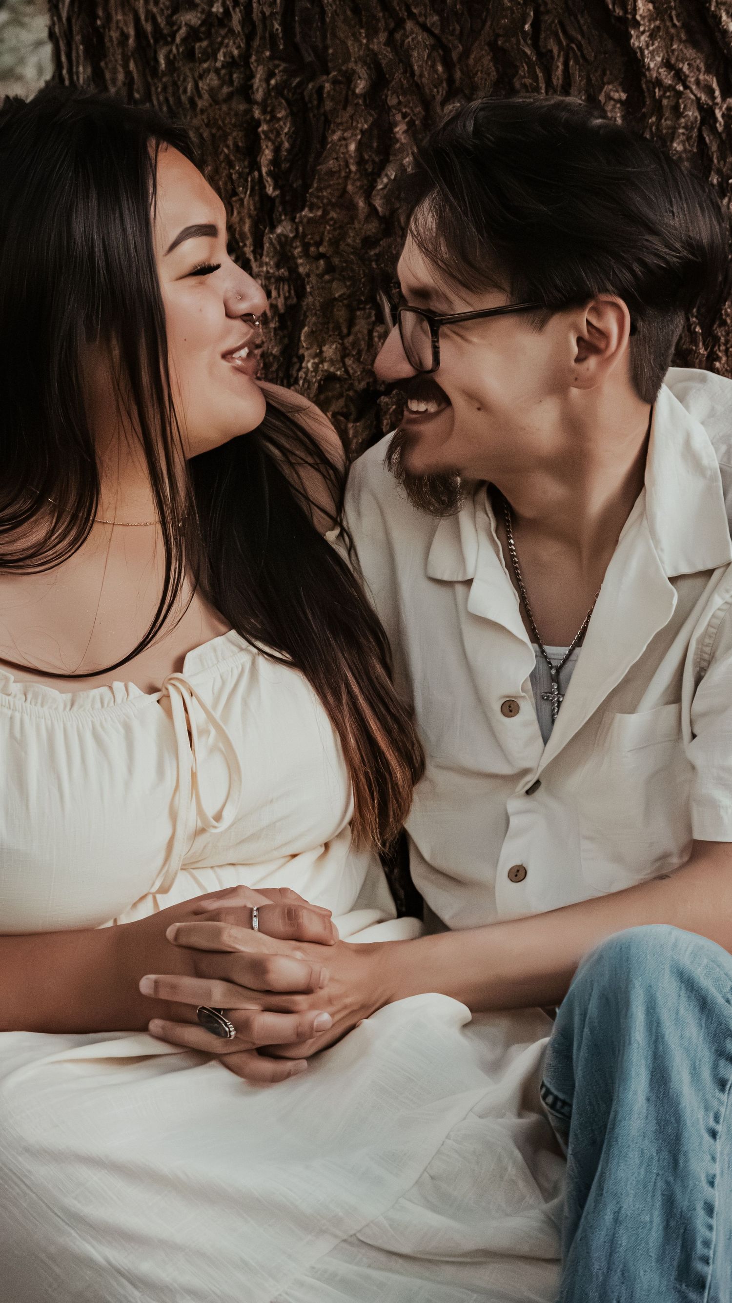 A happy couple smiles and embraces while sitting against a large tree trunk.