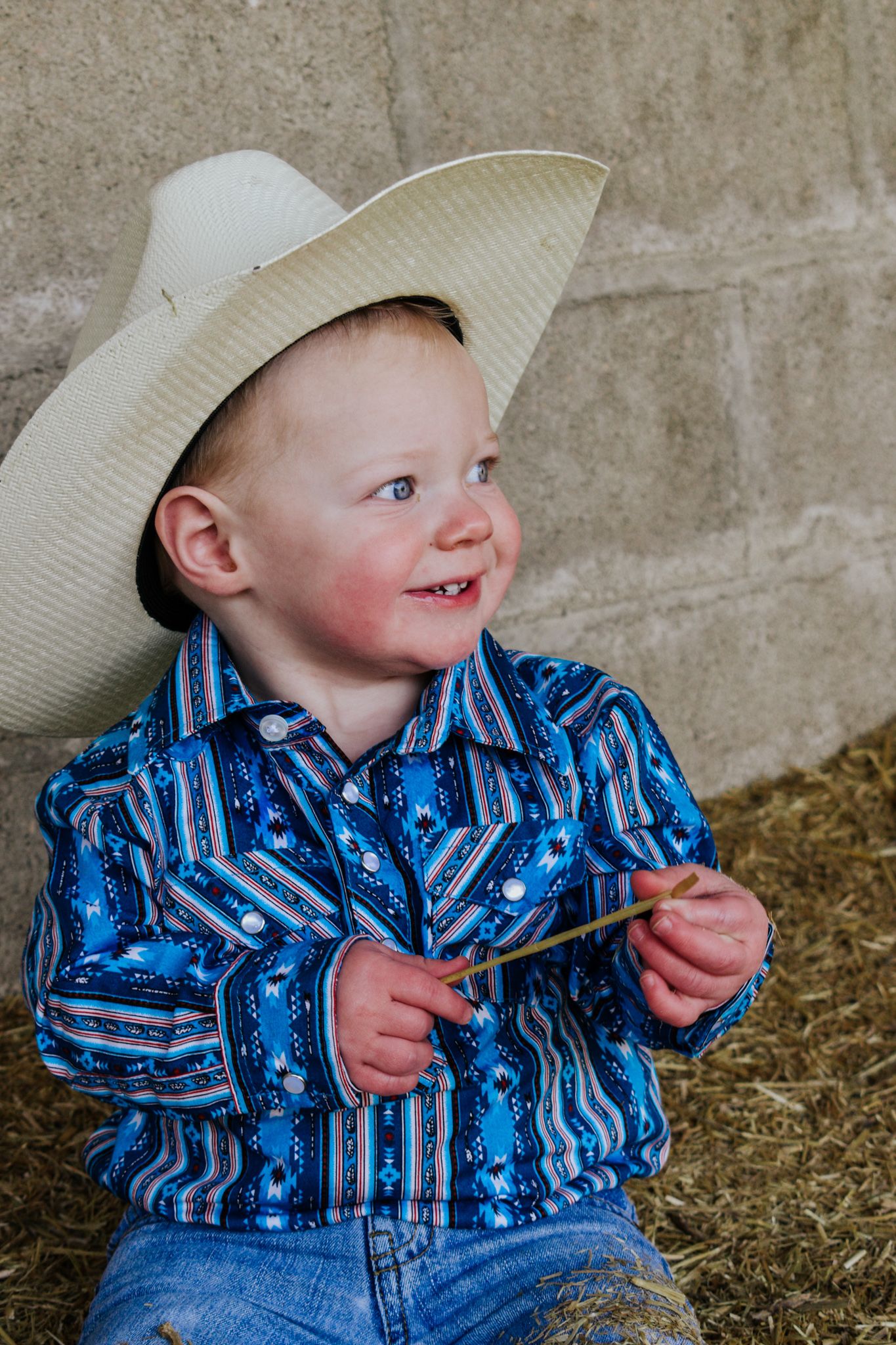 Smiling toddler boy wearing a white cowboy hat and blue western shirt sitting on hay. Family Photographer in Pflugerville, Tx