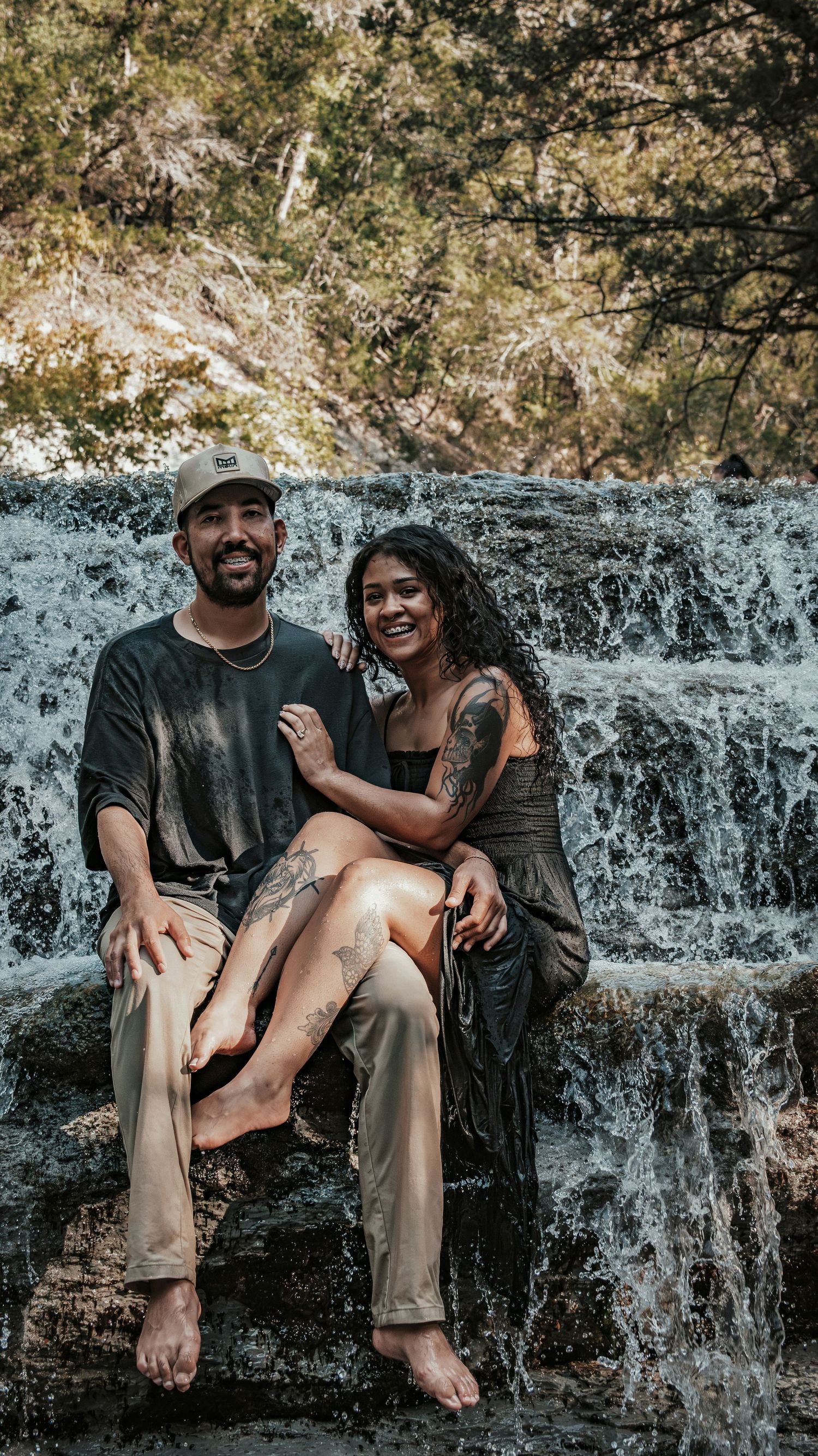 Smiling couple sitting together on rocks with a scenic waterfall backdrop outdoors for a couples session.