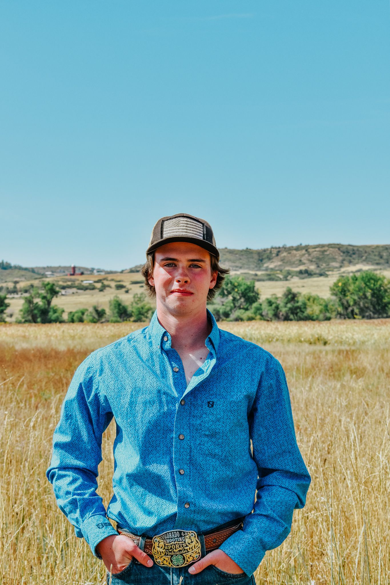 Young man in blue western shirt and trucker cap standing in dry grassland with hills in background for senior photos.