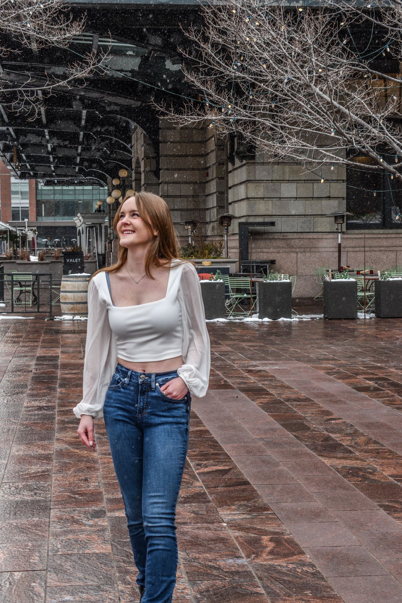 Cheerful young woman in white sheer-sleeve crop top and jeans strolling through snowy urban courtyard for senior photos.