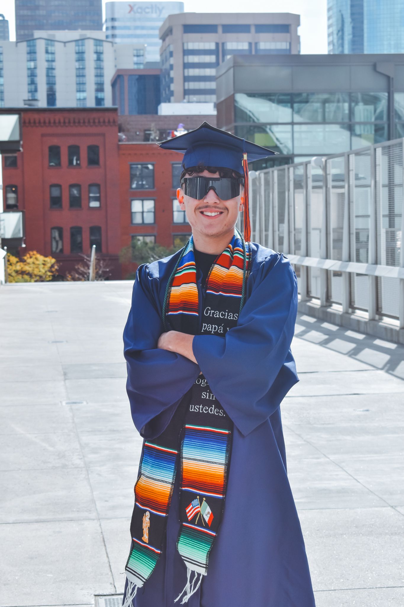 Smiling graduate in blue cap and gown with colorful stole poses in front of city skyline for senior photos.