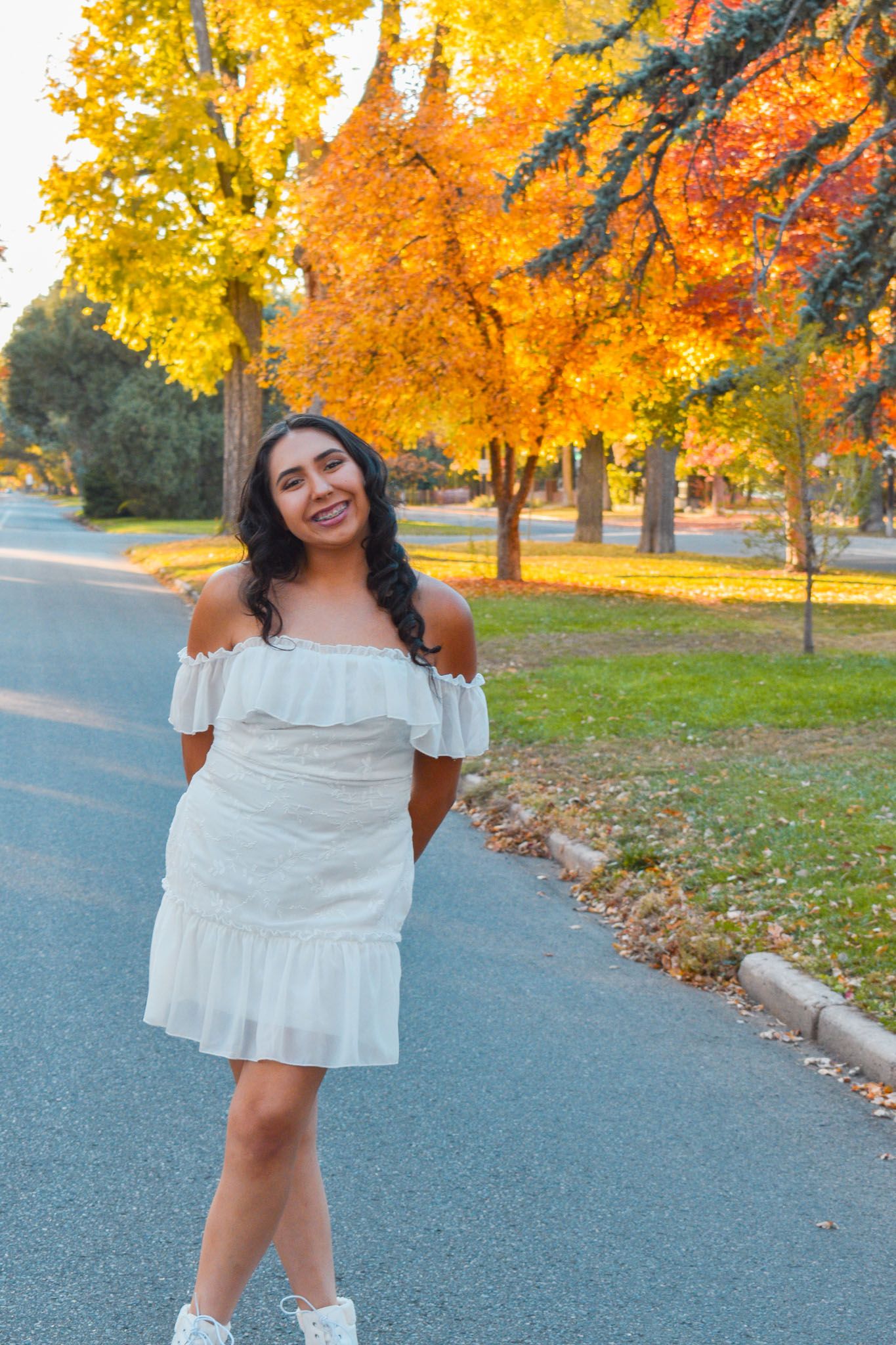 Young woman in white off-shoulder dress posing on tree-lined street with vibrant autumn foliage for senior photos.