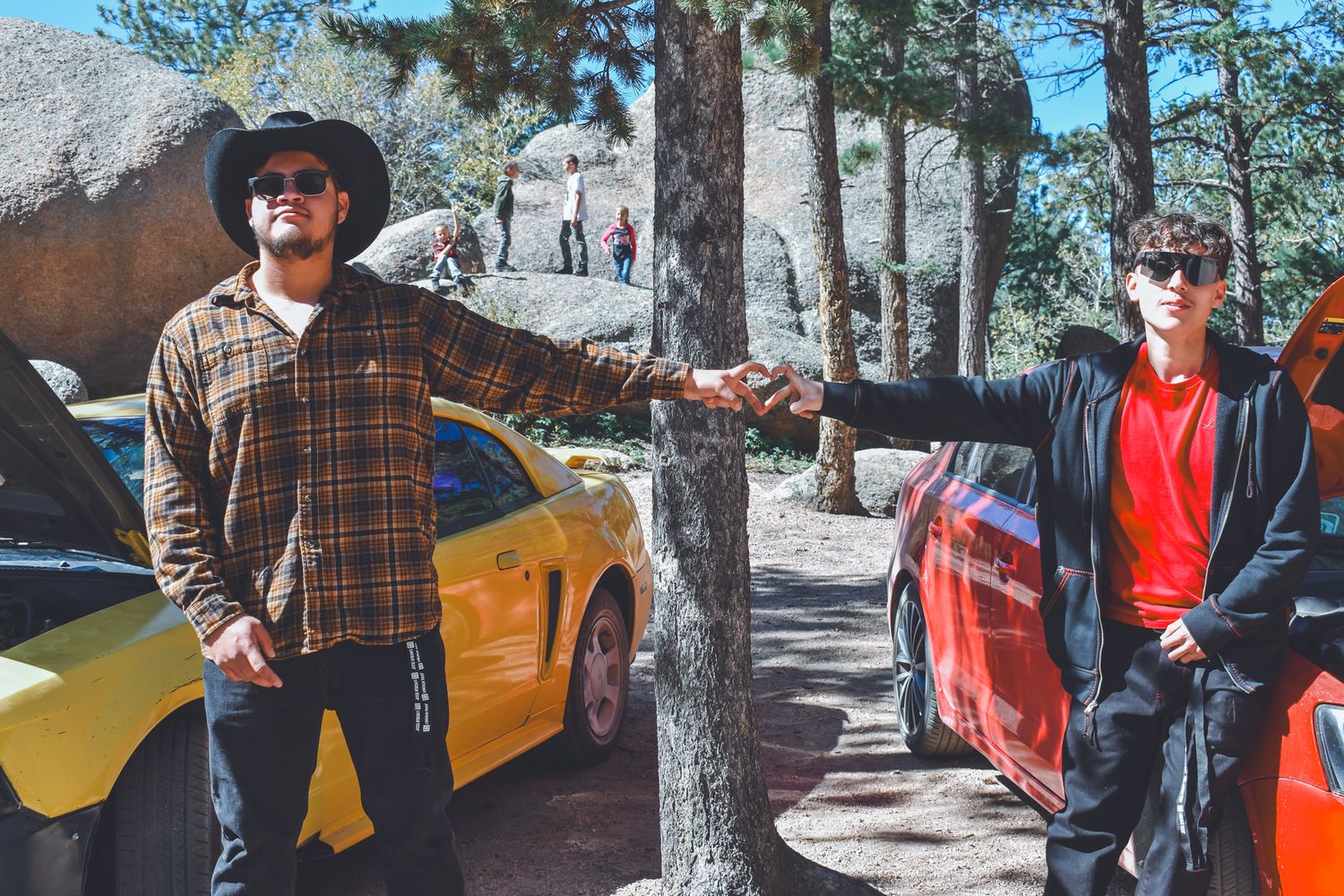 Two men fist bumping near colorful sports cars on a mountain road surrounded by pine trees for senior photos.