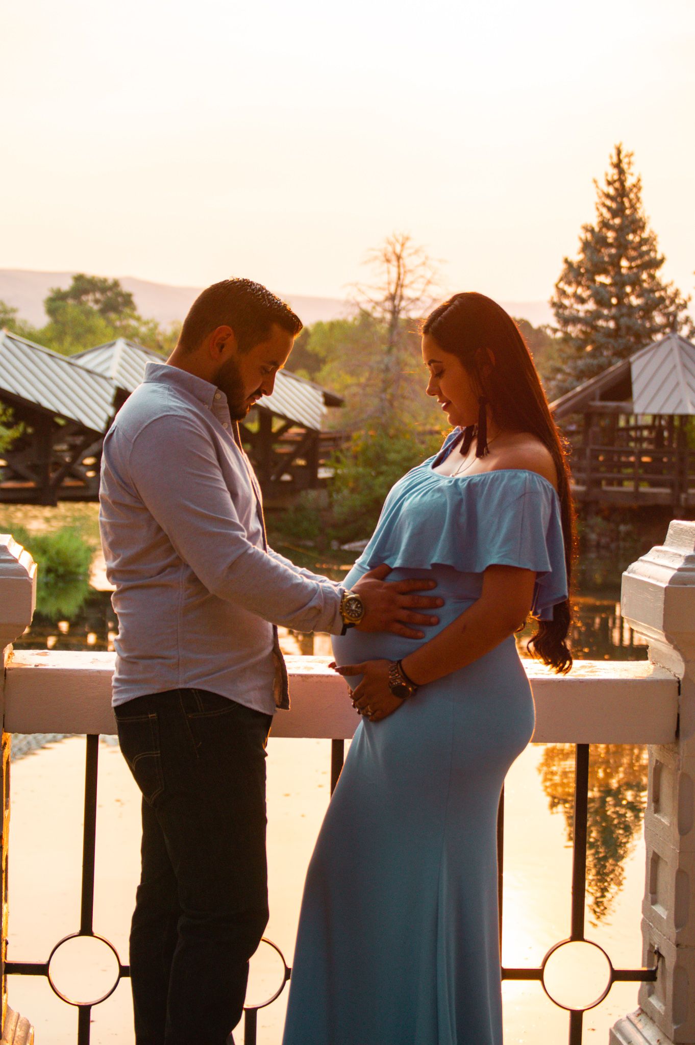 Couple tenderly holding pregnant belly on bridge at sunset golden hour.